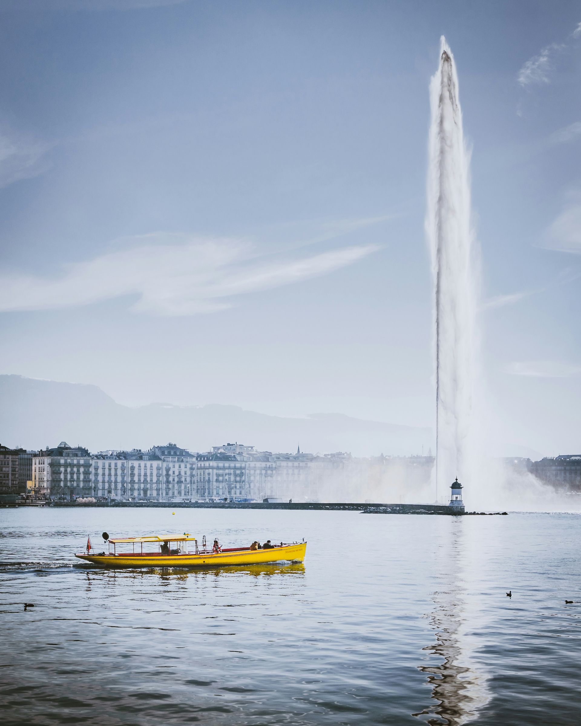 A yellow boat cruises on Geneva Lake near the Jet d'Eau fountain, with buildings in the background.
