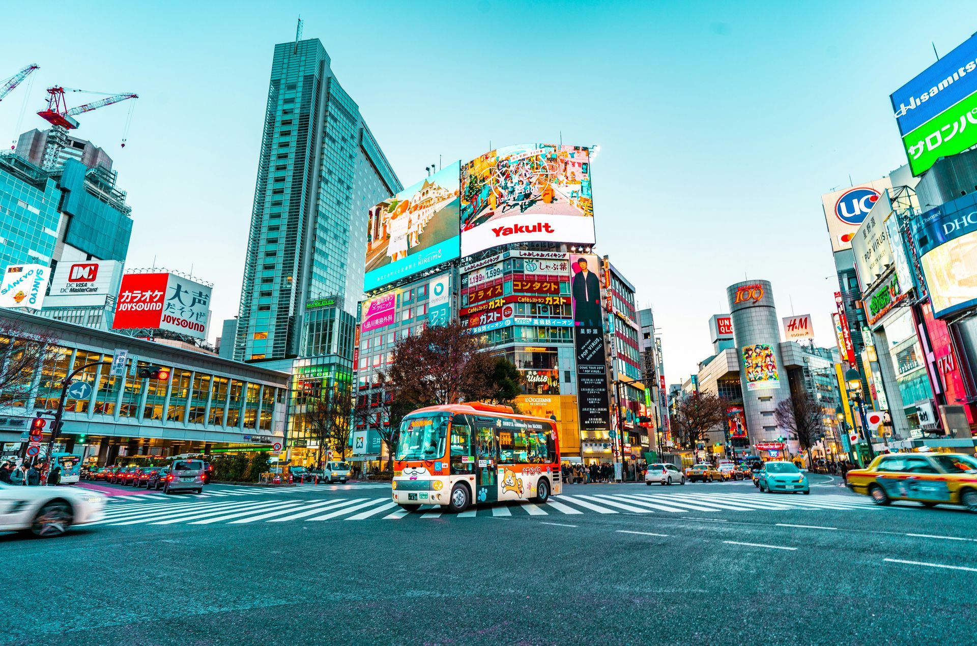 Busy Shibuya crossing in Tokyo, Japan, with multiple lit billboards and vehicle traffic on a crosswalk.