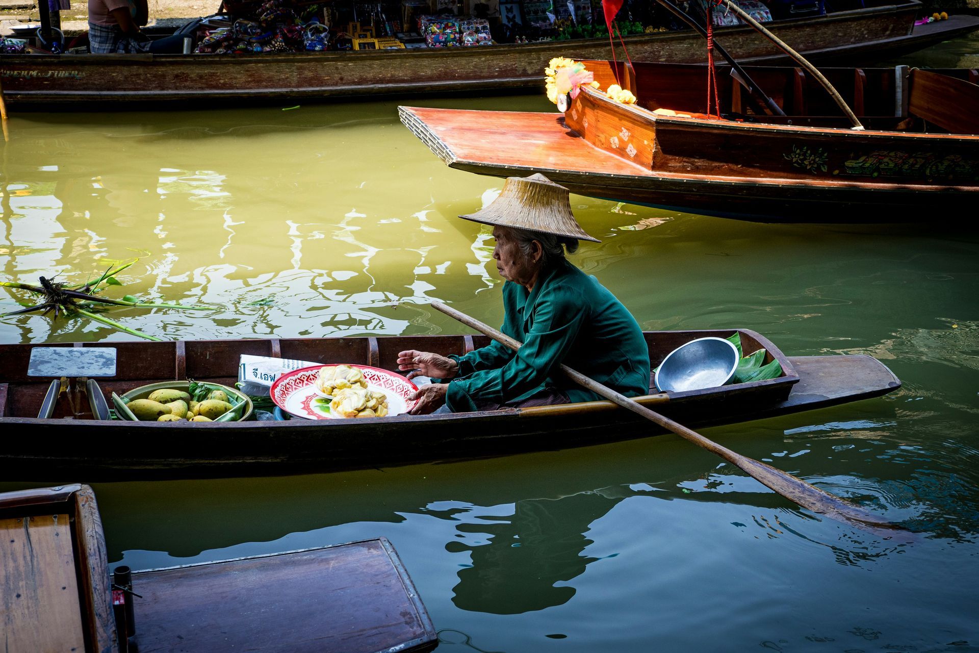 A woman in a green shirt is rowing a boat in the Damnoen Saduak river market in Thailand.