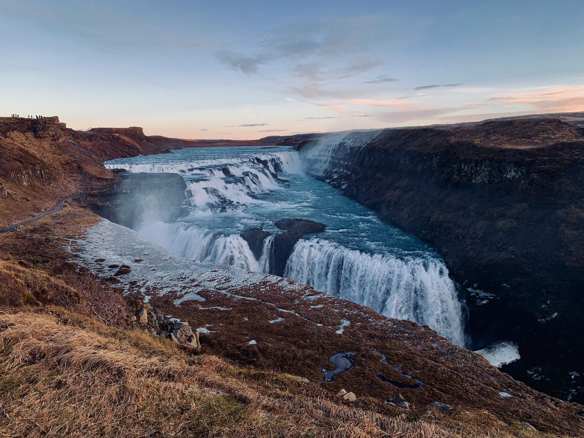Gullfoss Falls cascading into a canyon, with a blue-tinted river and brown cliffs under a pale sky in Iceland.