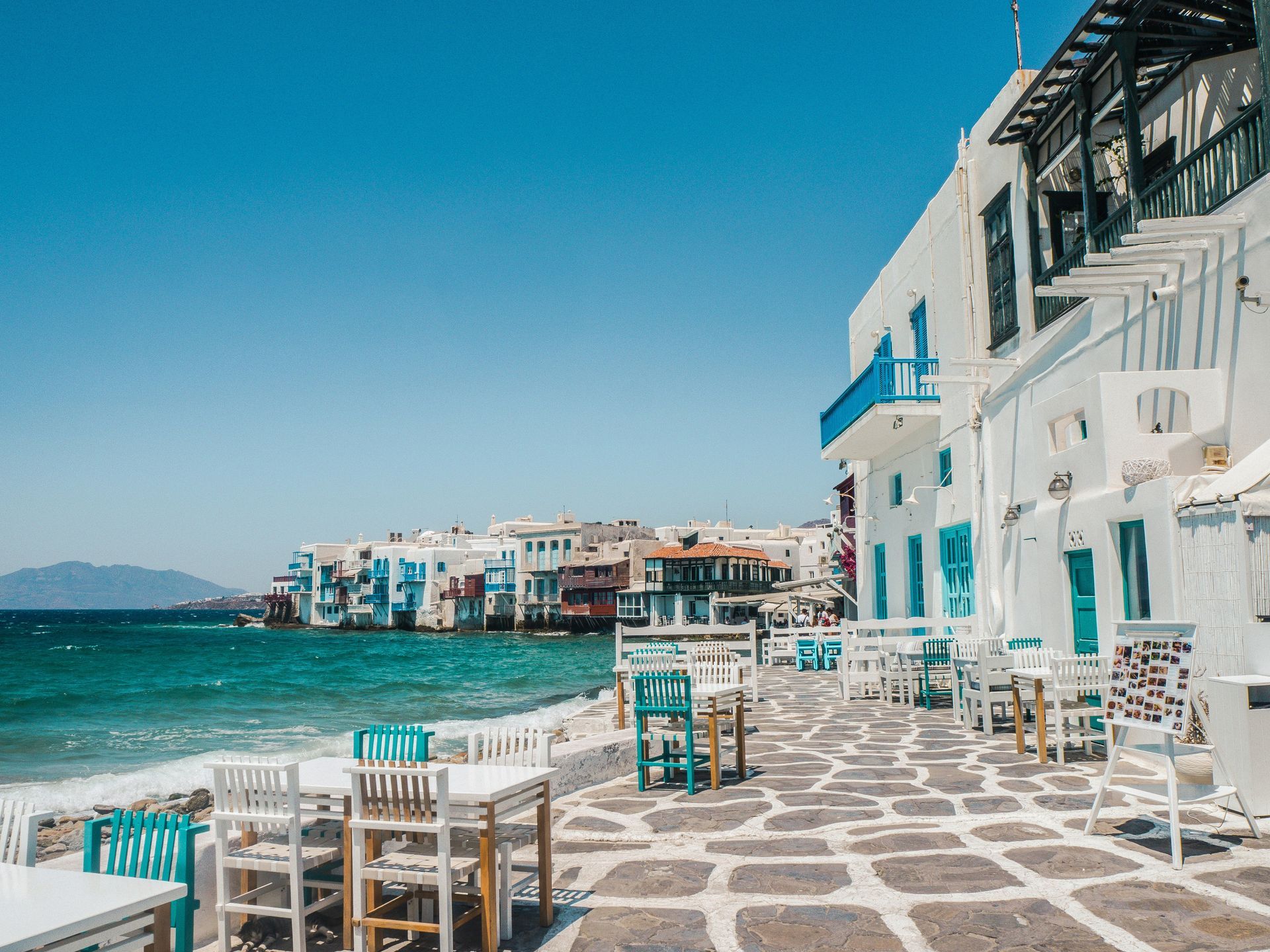 There are tables and chairs on the sidewalk next to the ocean in Mykonos, Greece.