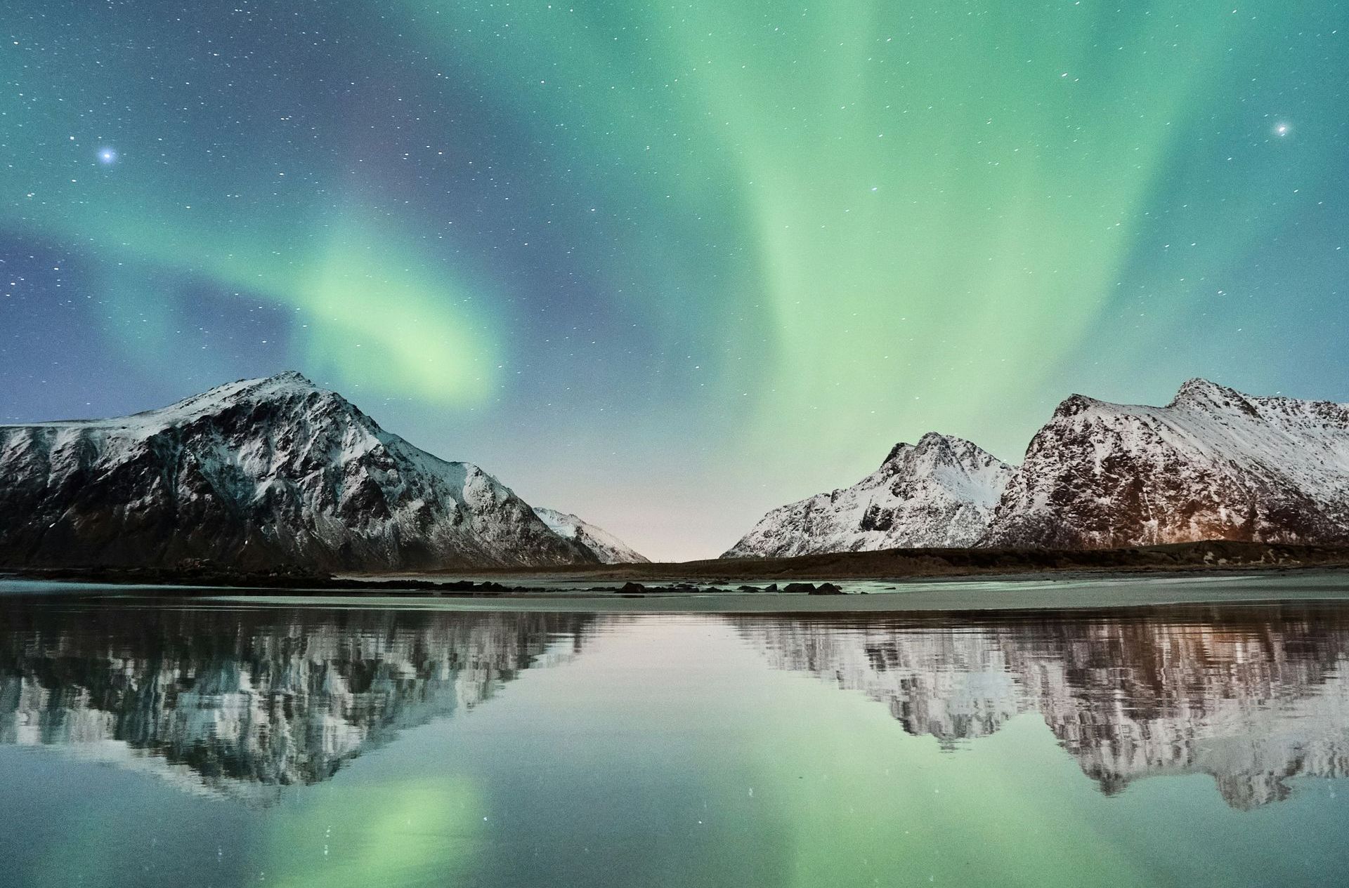 Aurora borealis over snow-capped mountains reflecting in calm water at night in Norway.