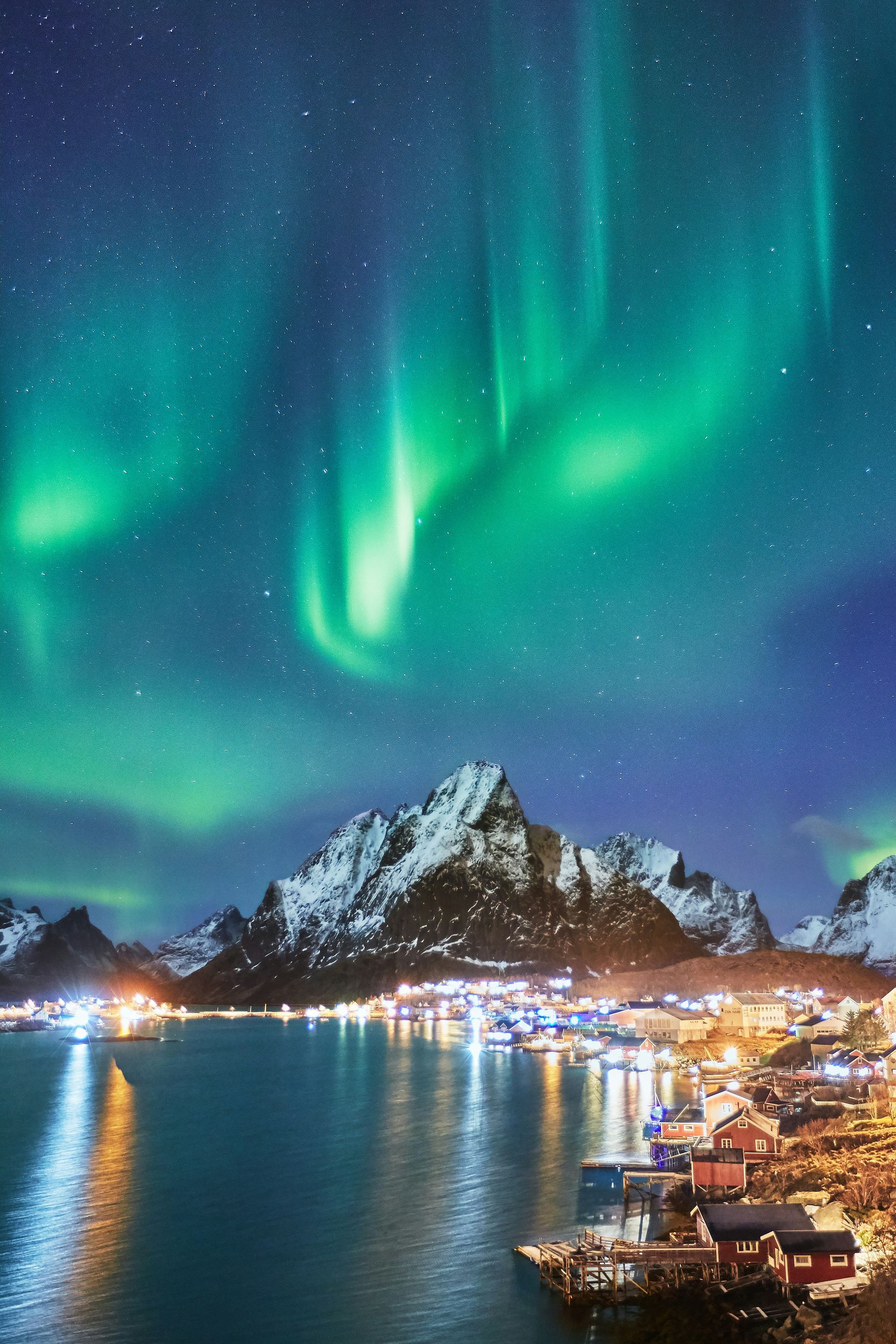 Aurora borealis over a snowy mountain range and the town of Lofoten, Norway on a fjord.
