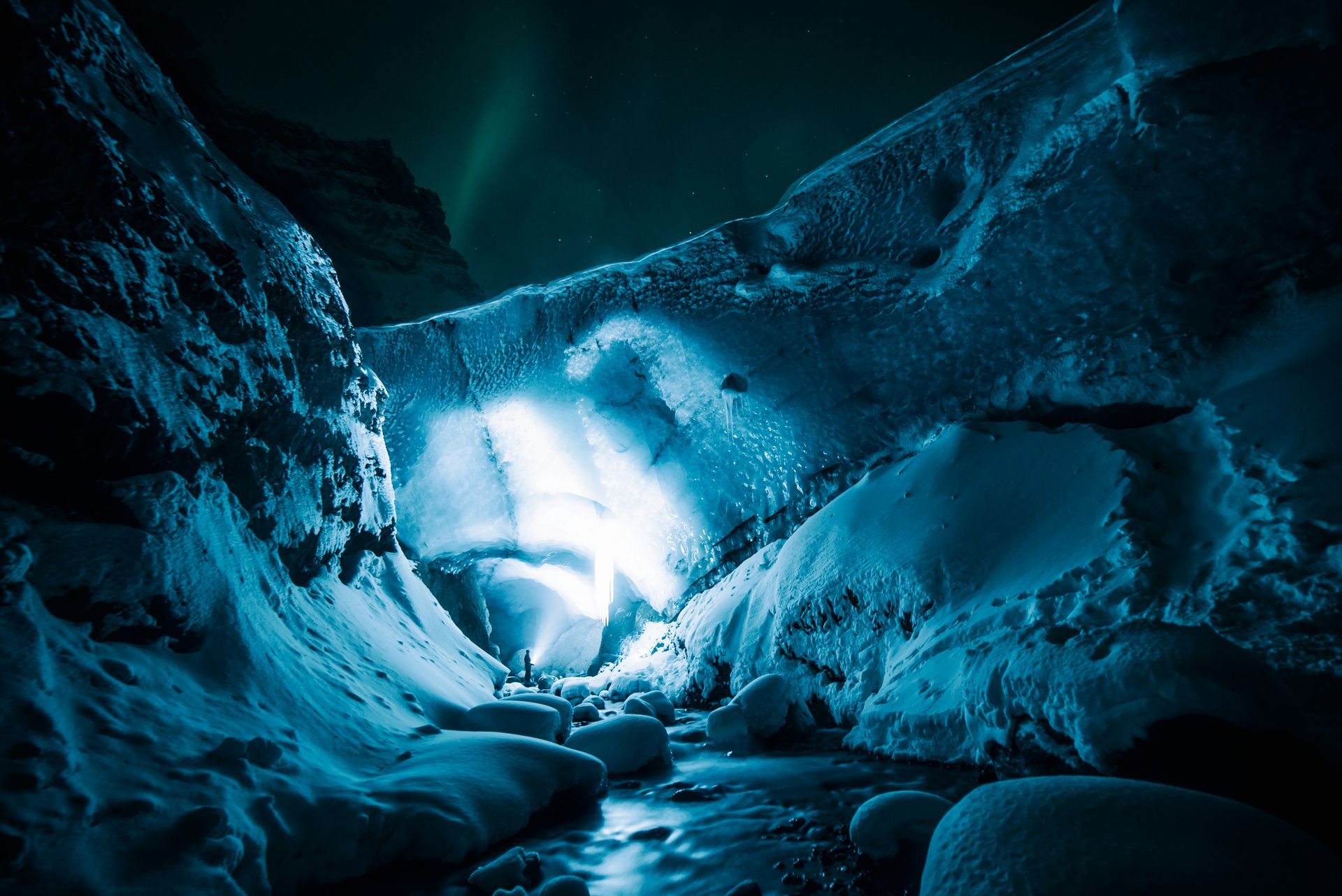 The Blue Ice Caves, Iceland, with a person standing, glowing white light, teal hues, aurora borealis.