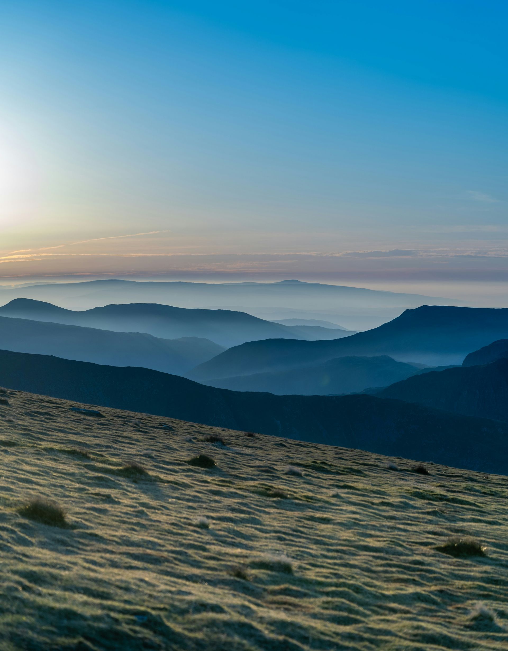 Mountains layered in shades of blue fade into the misty horizon under a clear, pale blue sky in the Lake District, England.