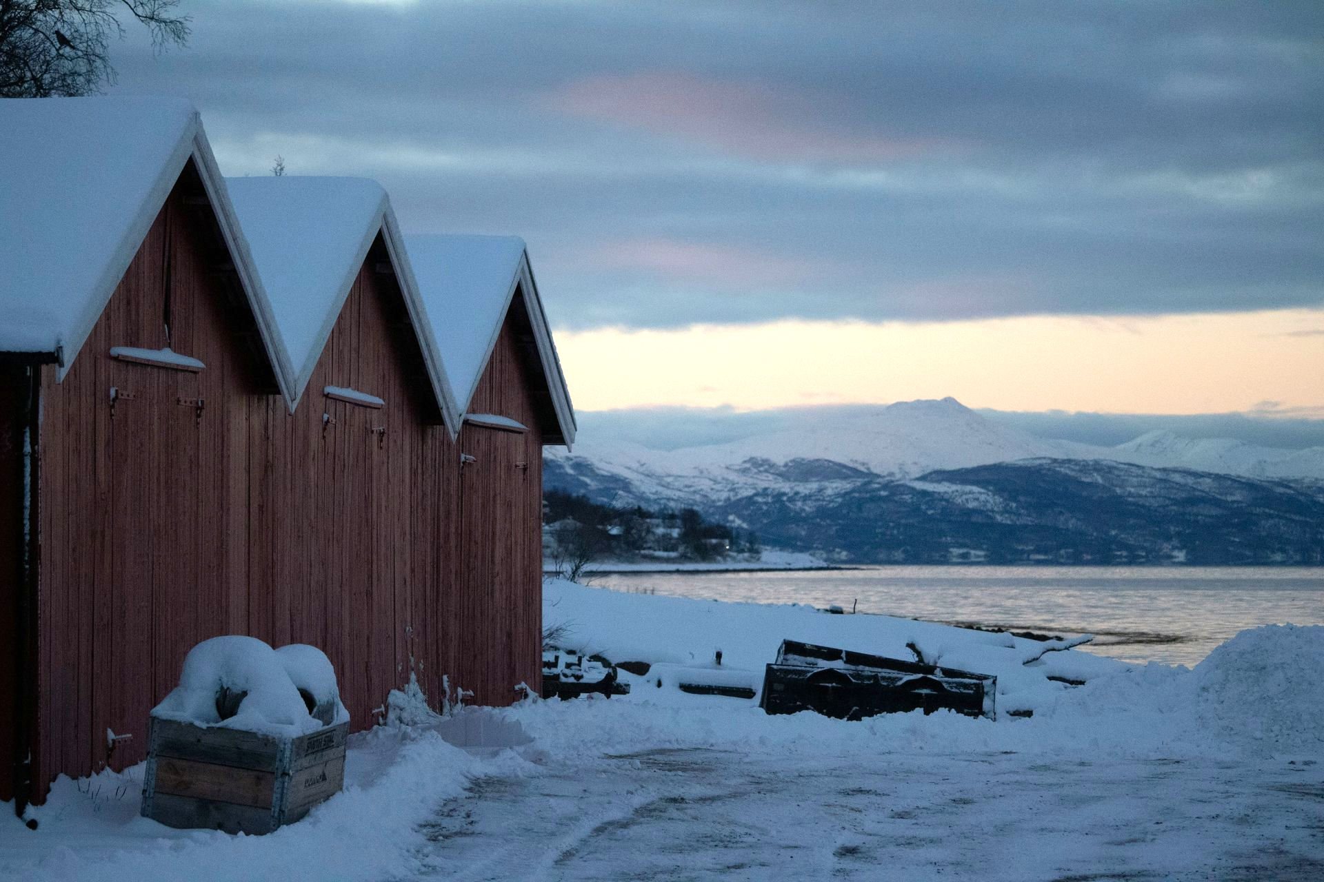 Red sheds with snow-covered roofs on a snowy shore, overlooking a lake and snow-capped mountains in Narvik, Norway.