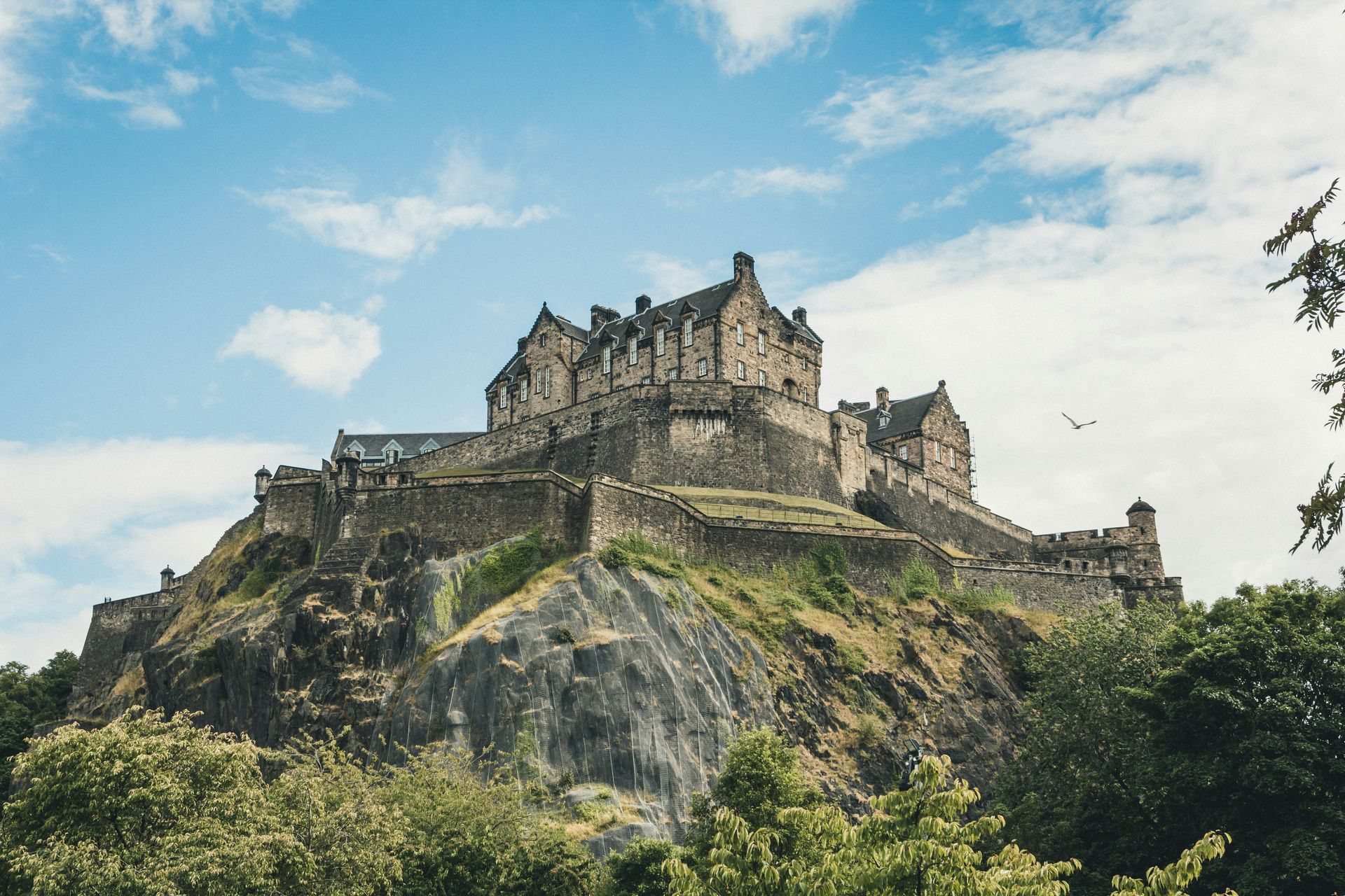 Edinburgh Castle atop a rocky hill against a blue sky with clouds, seen from below.