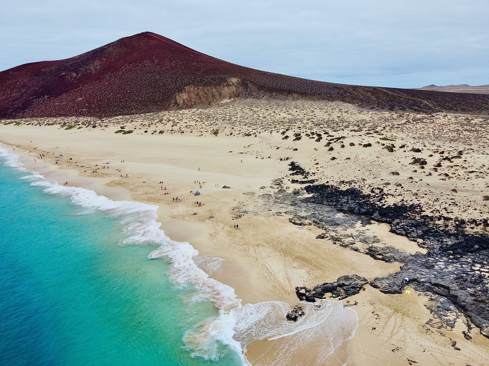 Beach with turquoise water, tan sand, and dark volcanic rock, backed by a dark red volcanic hill on the Canary Islands, Spain.