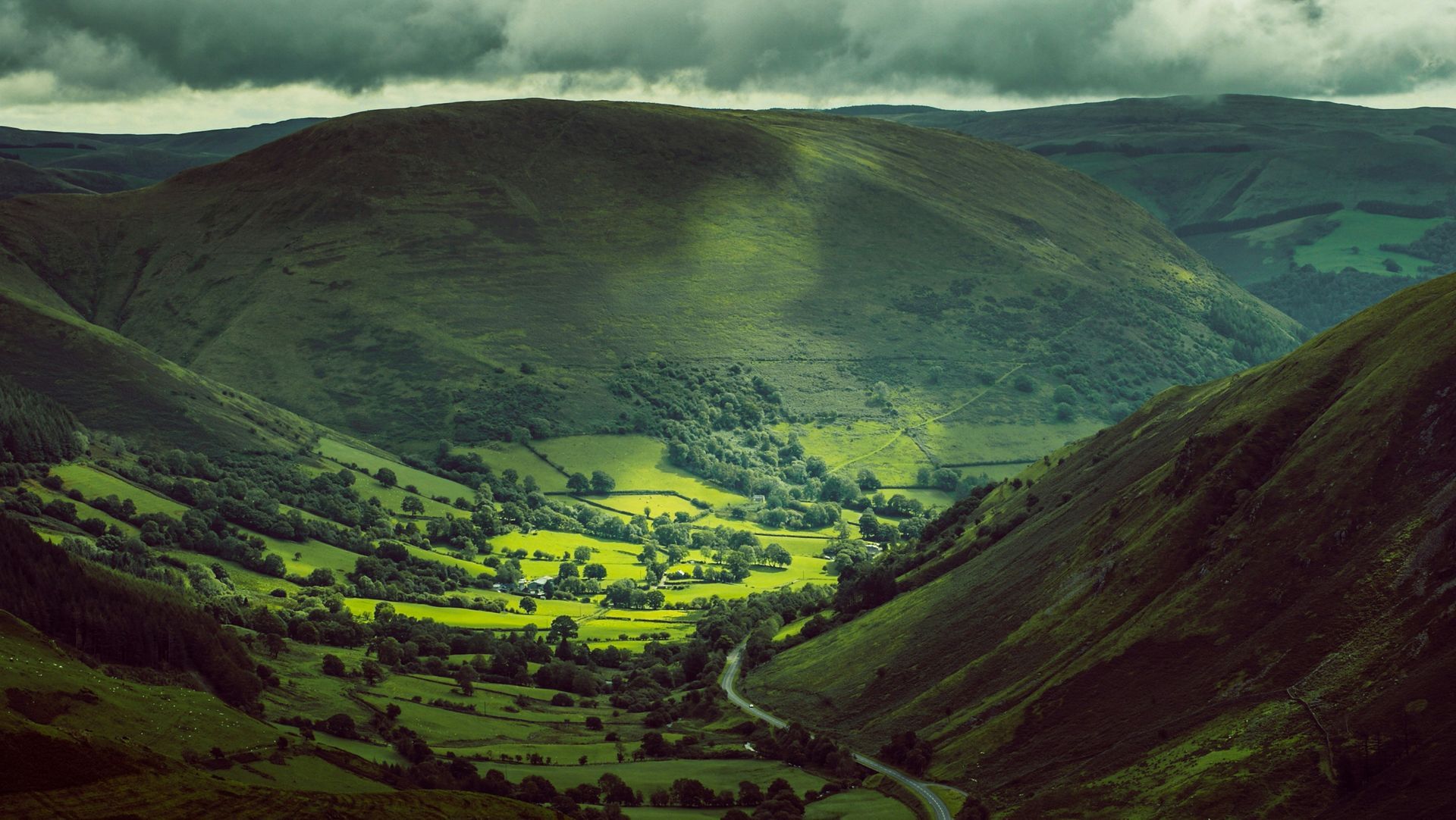Green valley with a winding road, nestled between mountains under a cloudy sky in Snowdonia, Wales.