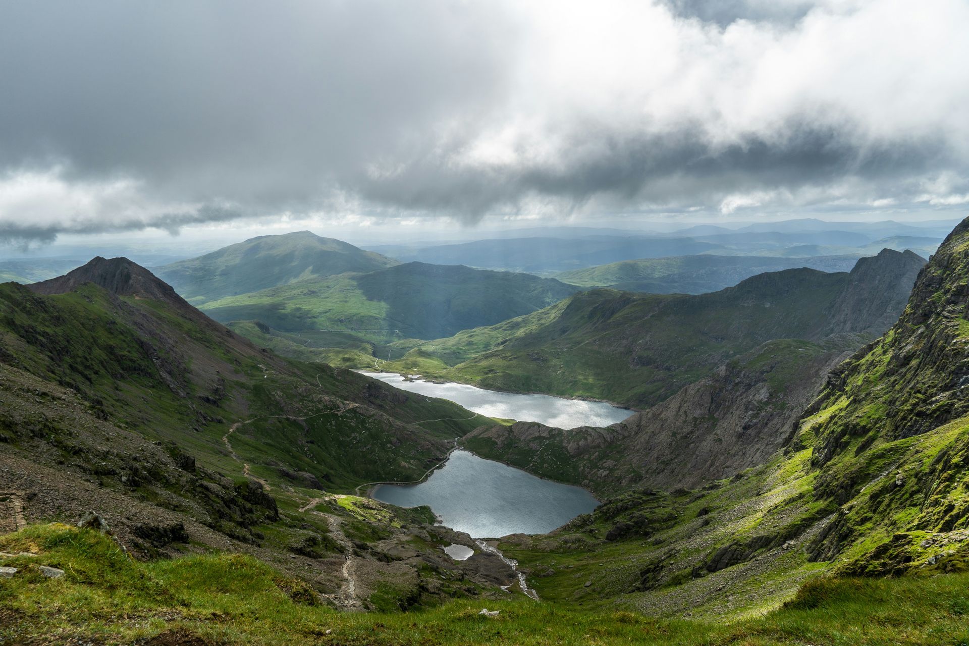 Mountains and two lakes viewed from a grassy slope under cloudy skies in Snowdonia, Wales.