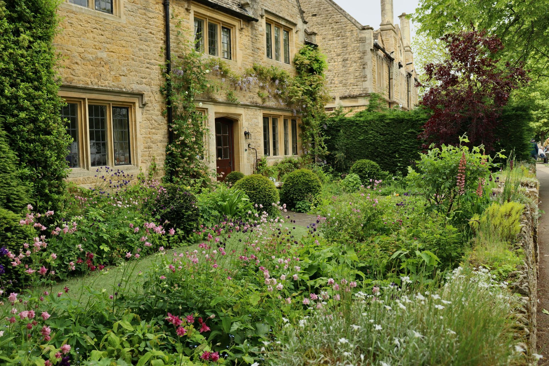 Cotswold stone cottage with a lush garden. Green bushes, colorful flowers, and a brown door in England.