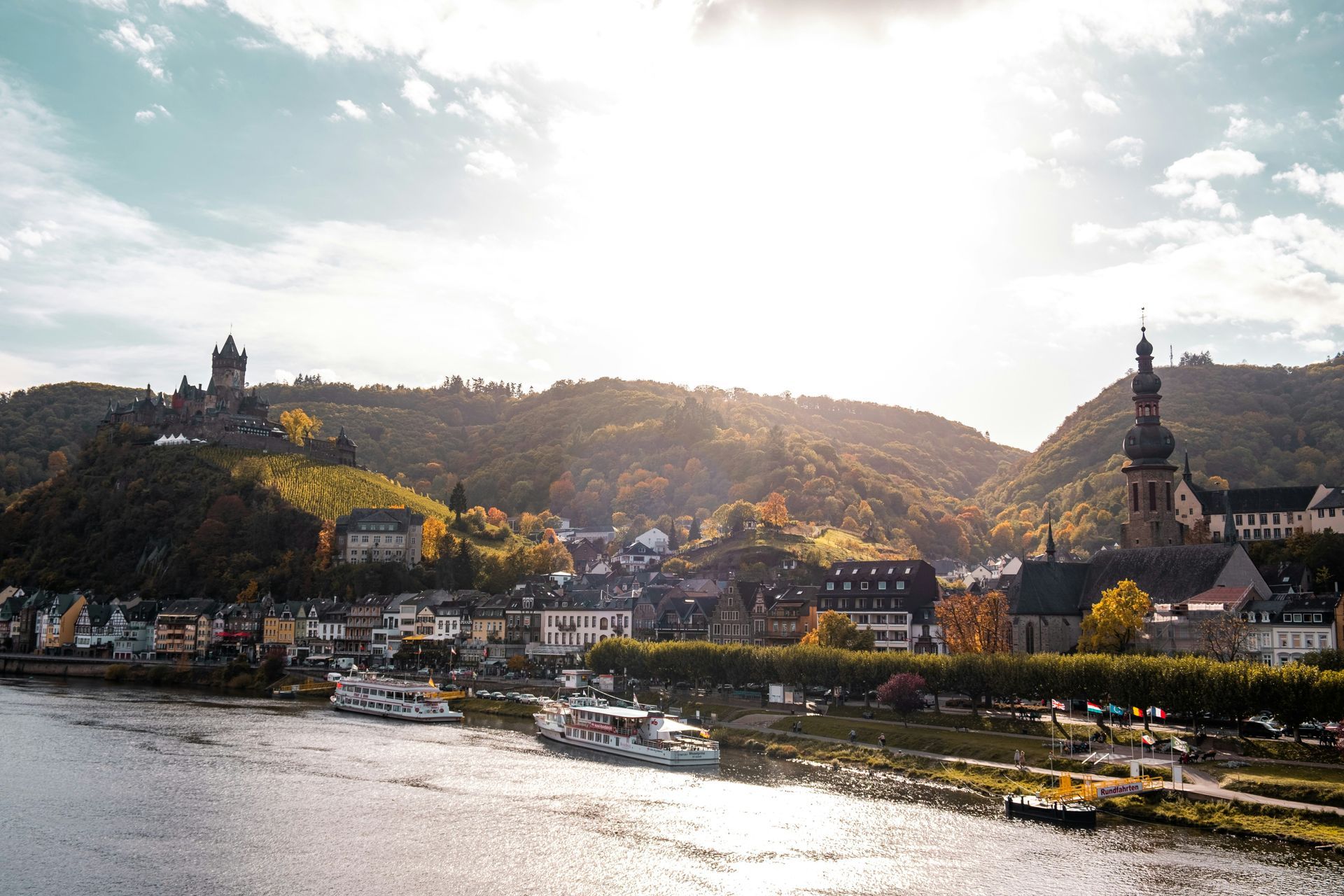 Riverfront town of Cochem in Germany with castle on a hill, church, boats, and fall foliage under a bright sky.