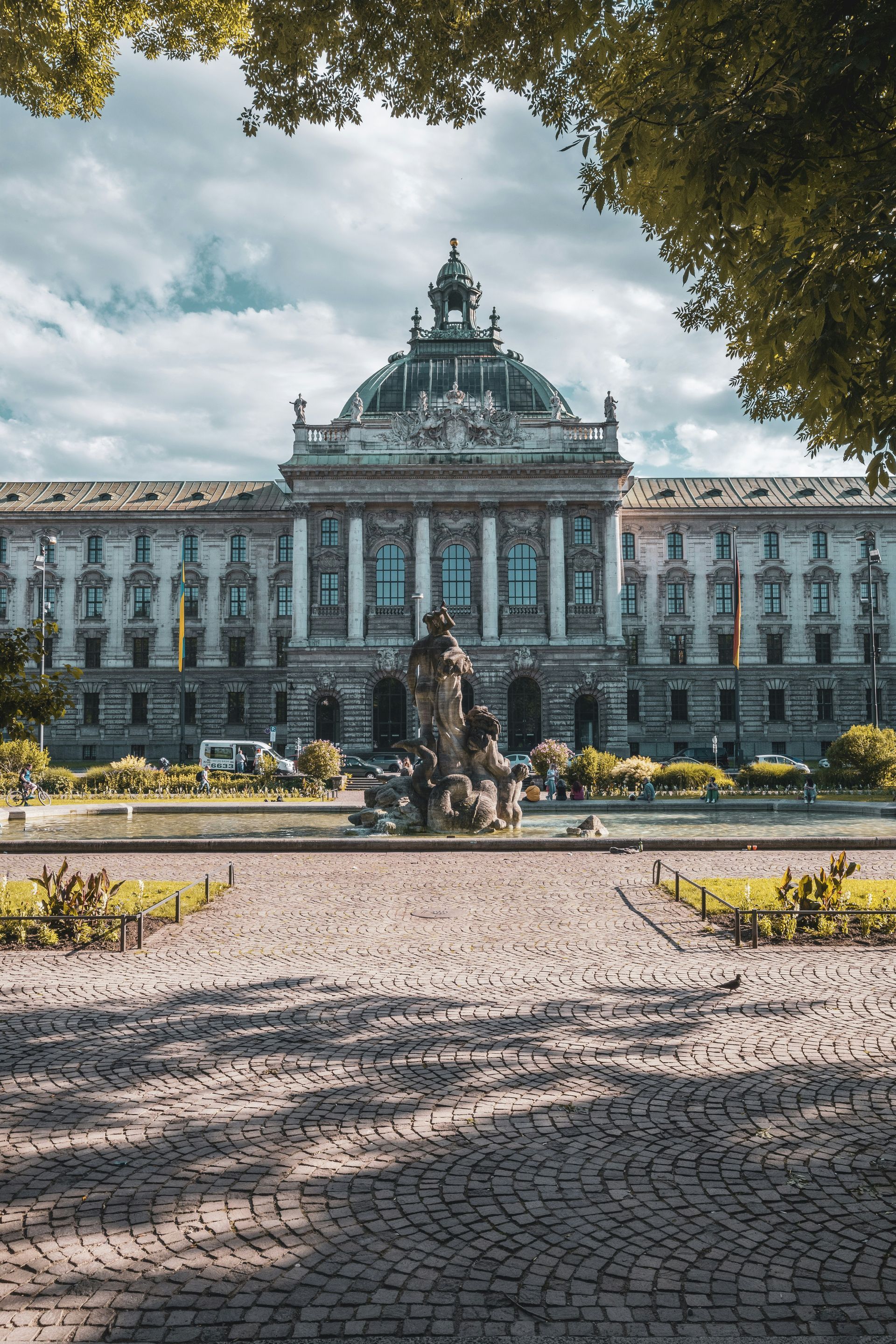 Ornate building with dome and fountain, viewed across cobblestone path in Munich, Germany.