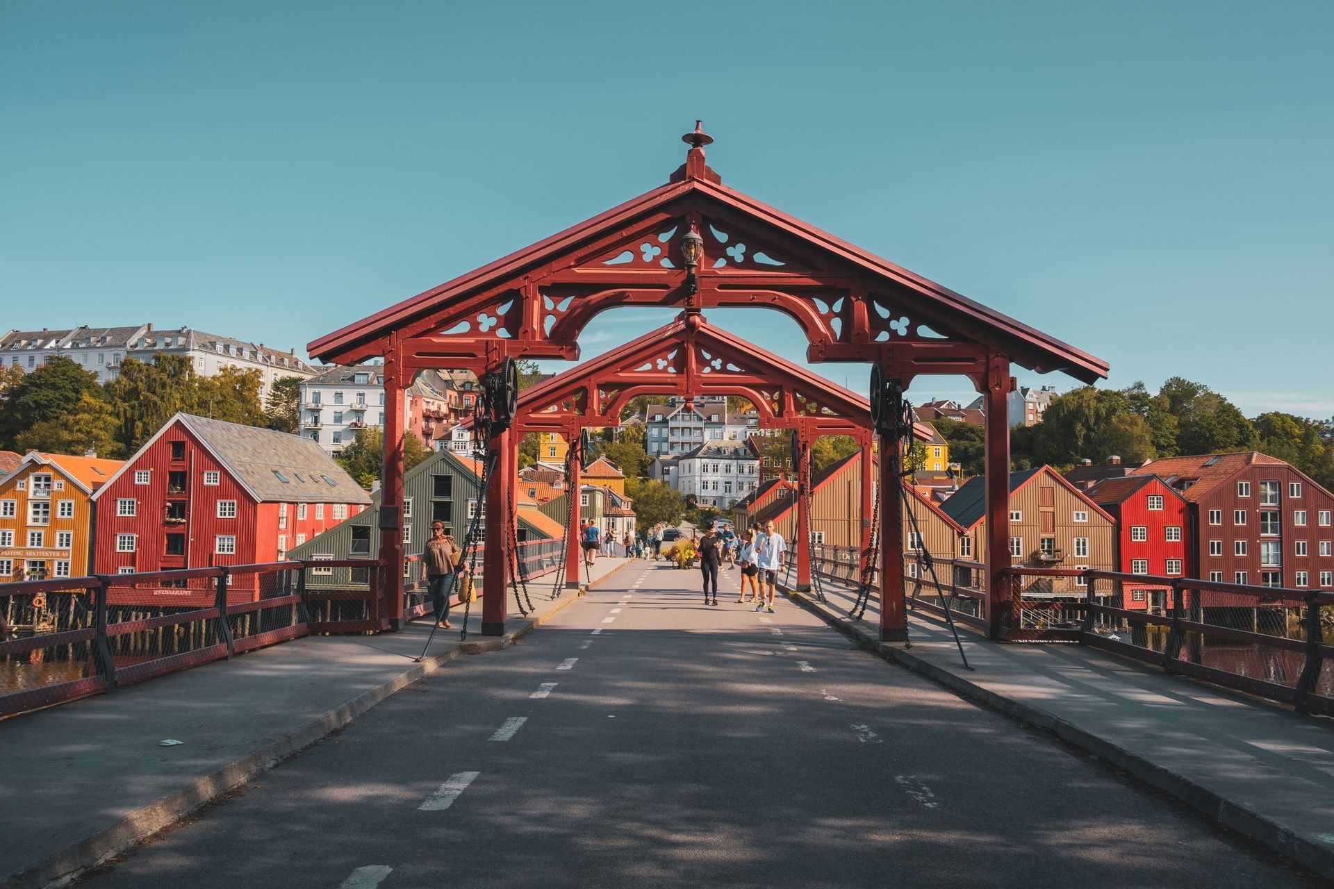 Red wooden bridge over a street in Trondheim, Norway, with colorful buildings and people walking.