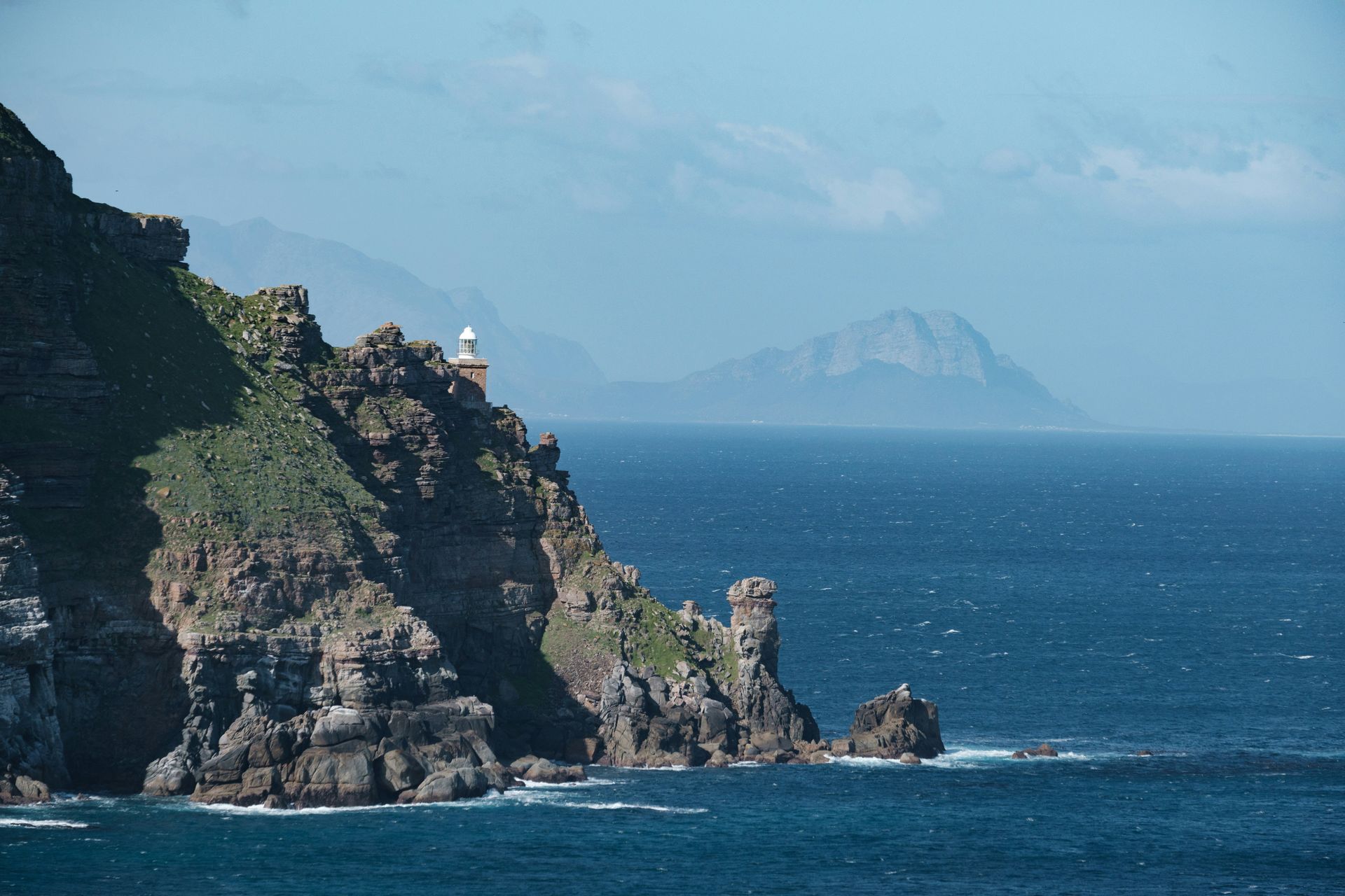 Rocky coastline with lighthouse, blue ocean, and distant mountains on the Cape Peninsula, South Africa.