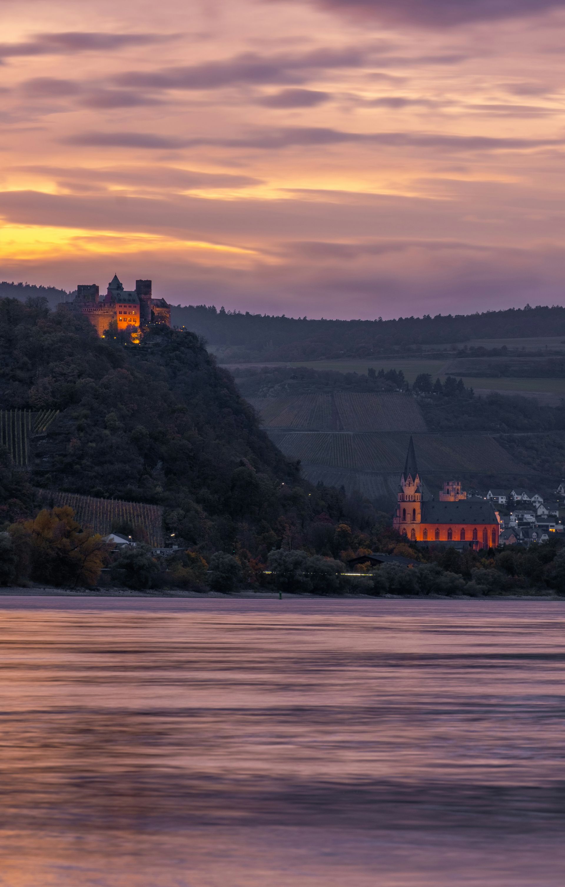 Twilight view of a castle on a hillside above the Rhine River Gorge, with a church in the distance.
