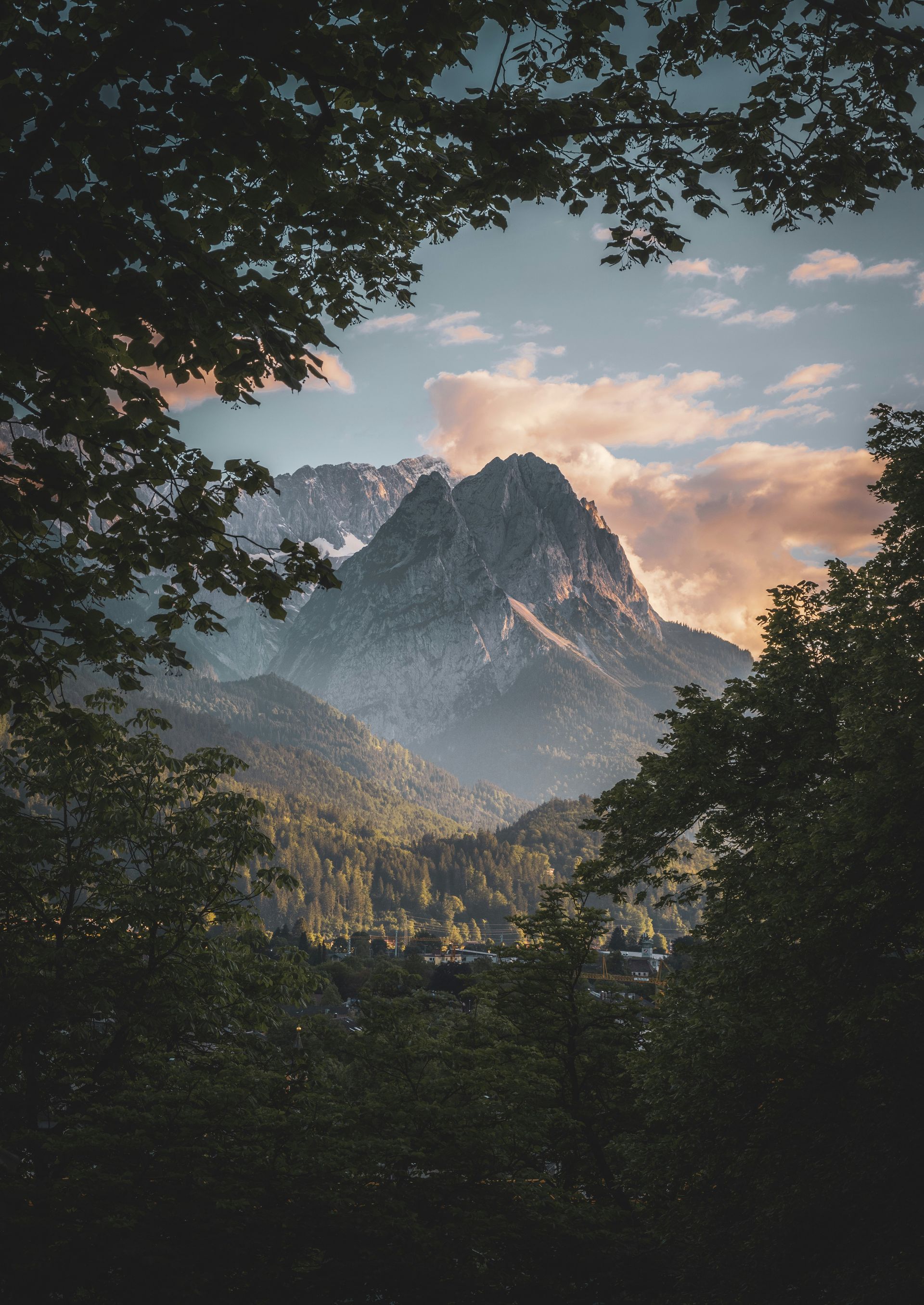 Mountain framed by tree branches; golden sunset illuminates peak and surrounding forest in Garmisch-Partenkirchen, Germany.