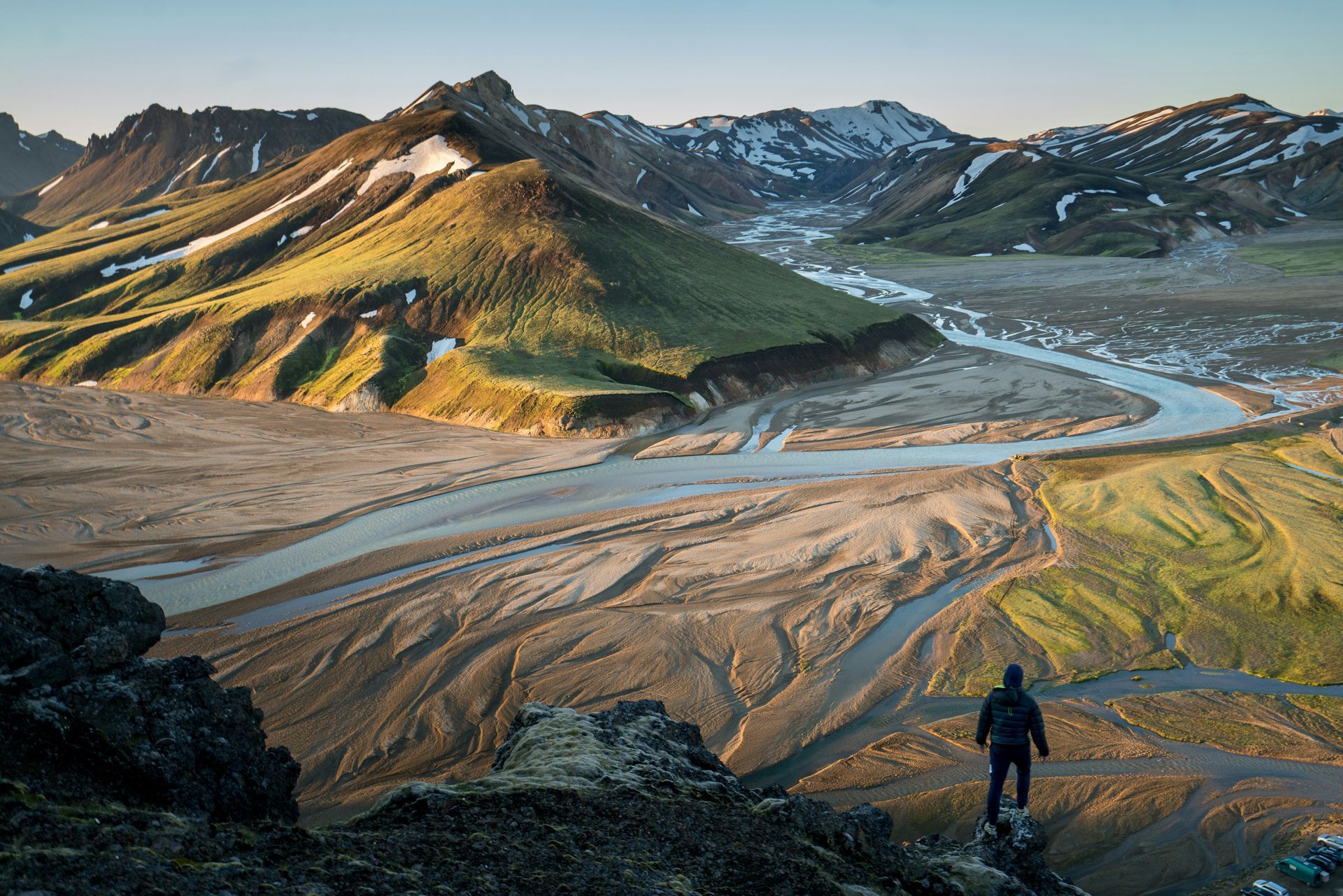 Man overlooking a vast river valley surrounded by green mountains. Sunlight illuminates the landscape in Landmannalaugar, Iceland.