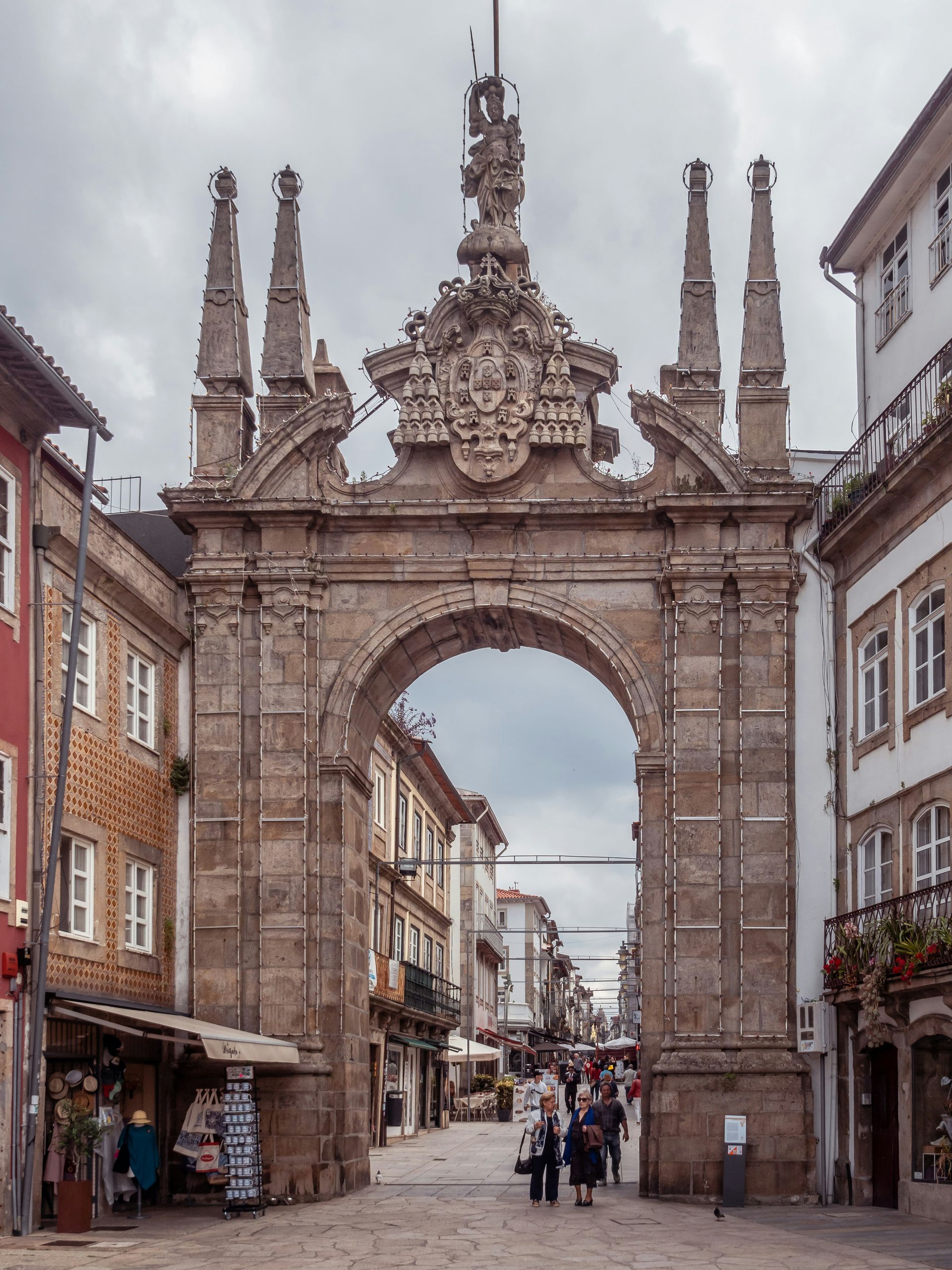 Stone archway with ornate carvings, pedestrians walking on cobblestone street, buildings on either side in Braga, Portugal.