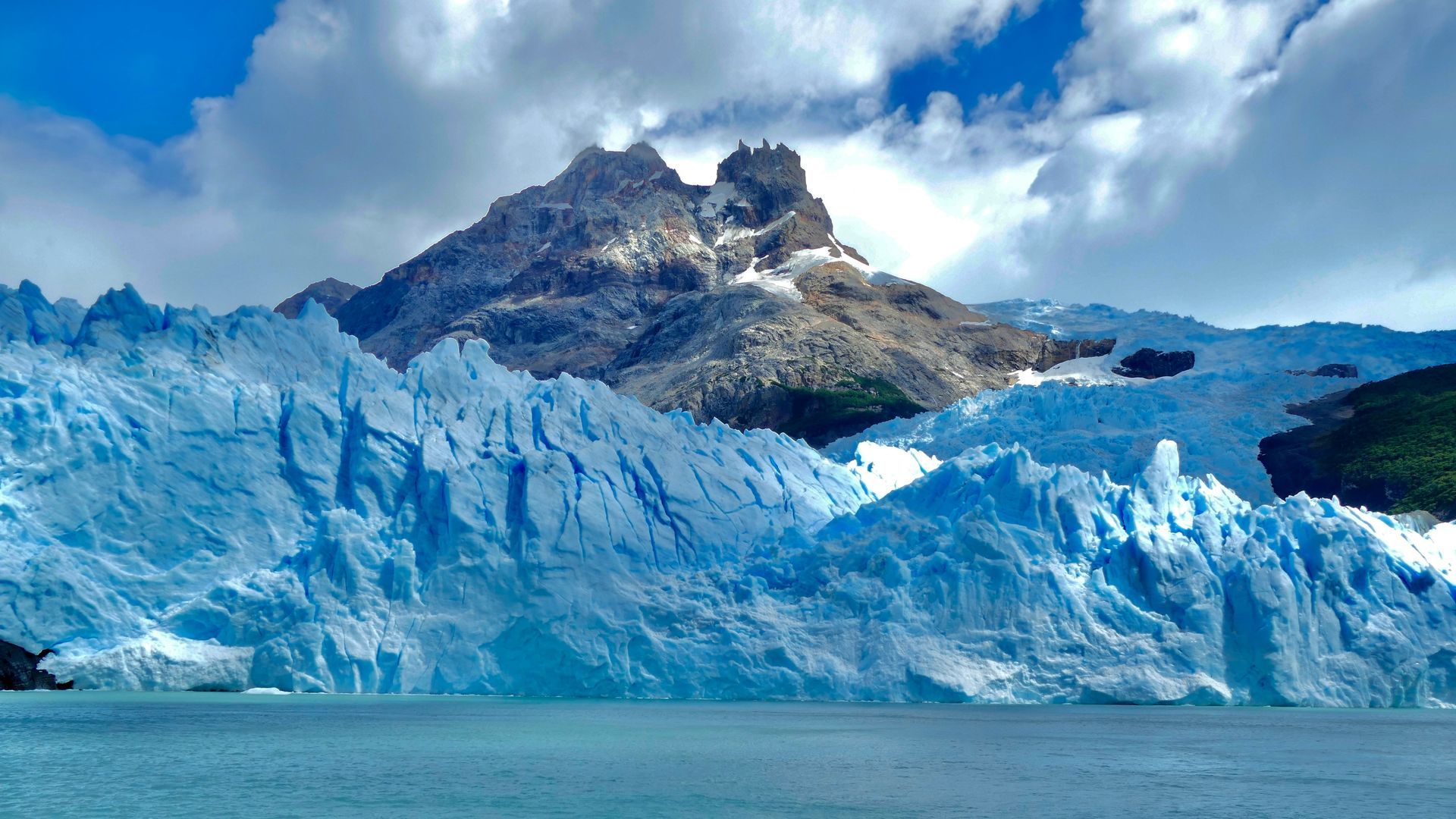 Blue glacier with jagged peaks and a mountain under a cloudy sky at Parque Nacional Los Glaciares in El Calafate, Argentina.