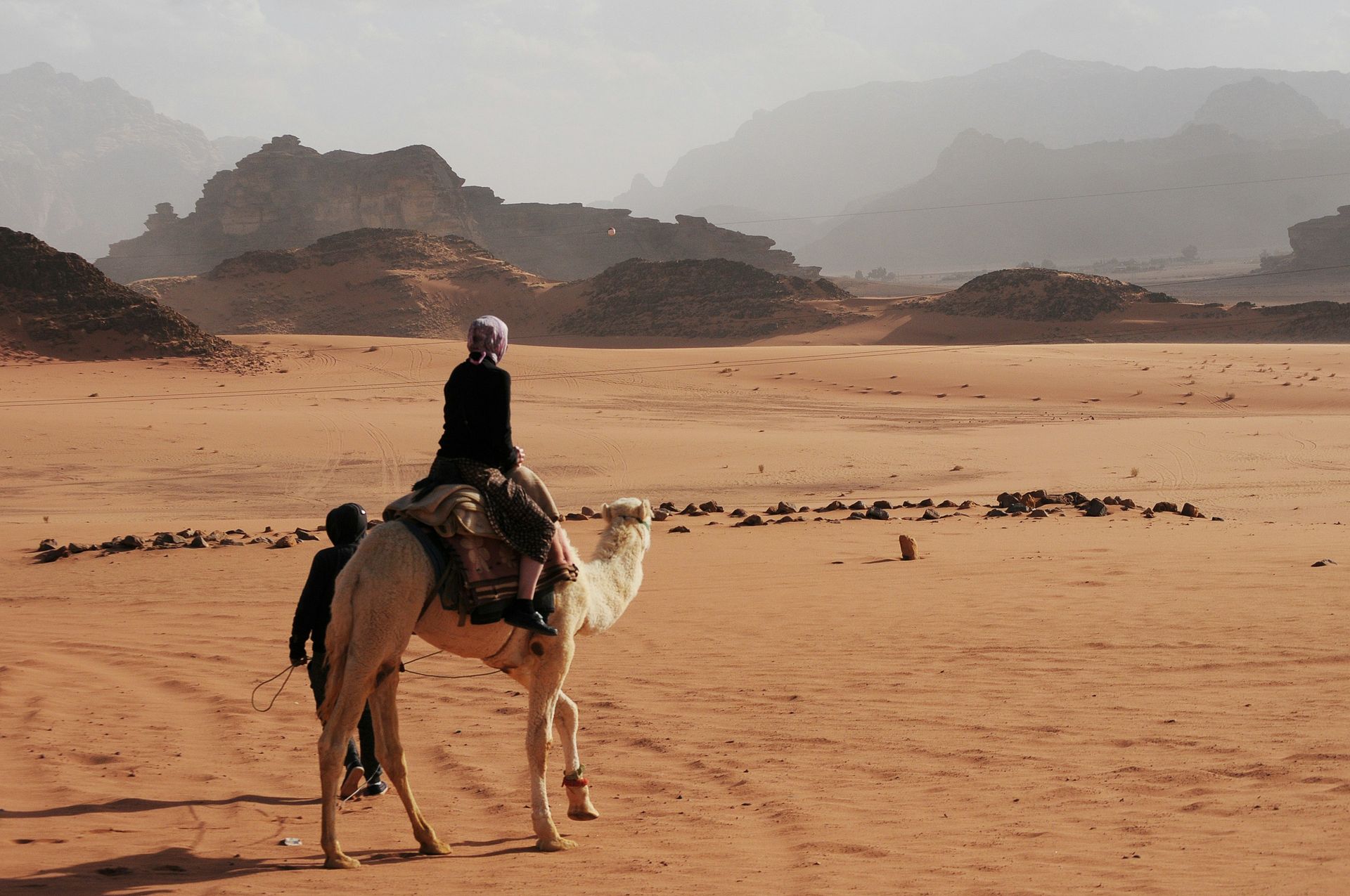 Person riding a camel in a desert landscape with hazy mountains in the distance in Wadi Rum, Jordan.
