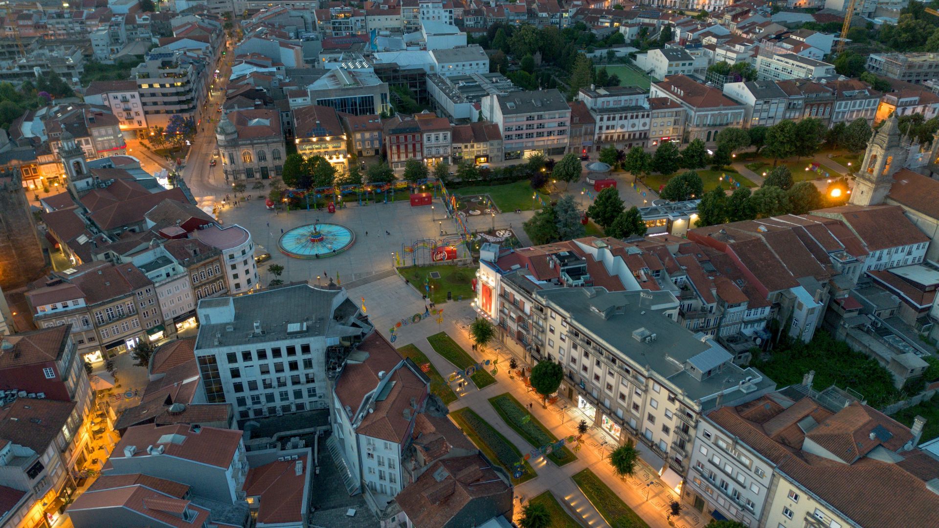 Aerial view of Braga, Portugal's city square at dusk; buildings with orange roofs surround a plaza, illuminated by lights.