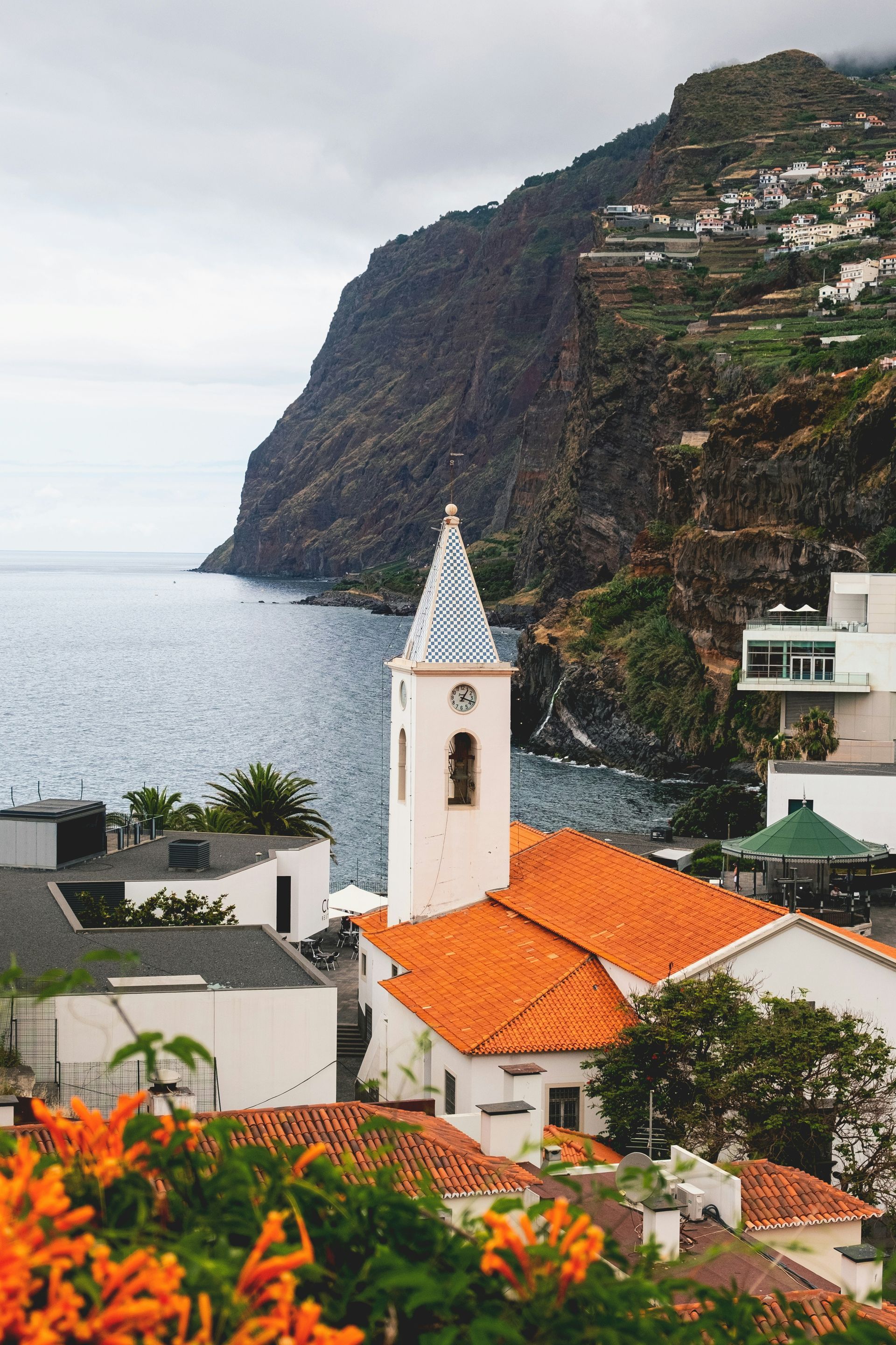 White church with orange roof and steeple by the coast, near a cliffside village on the island of Madeira, Portugal.