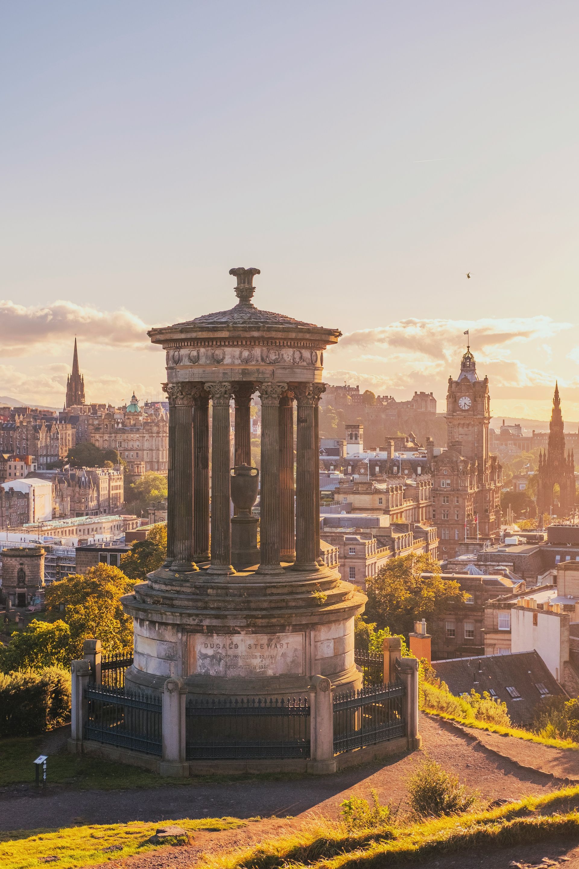 Dugald Stewart Monument in Edinburgh, Scotland. Golden hour sunlight bathes the neoclassical structure and cityscape.