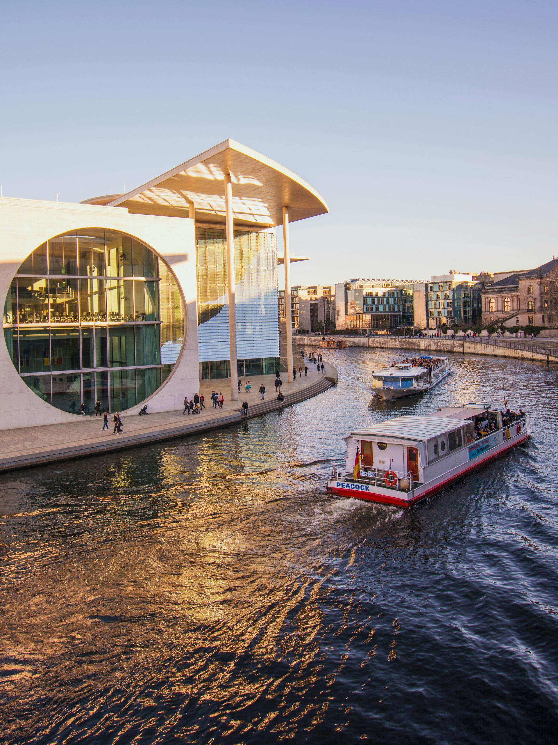 Boats on a river in Berlin, Germany, passing a modern building with large circular windows and a unique roof.