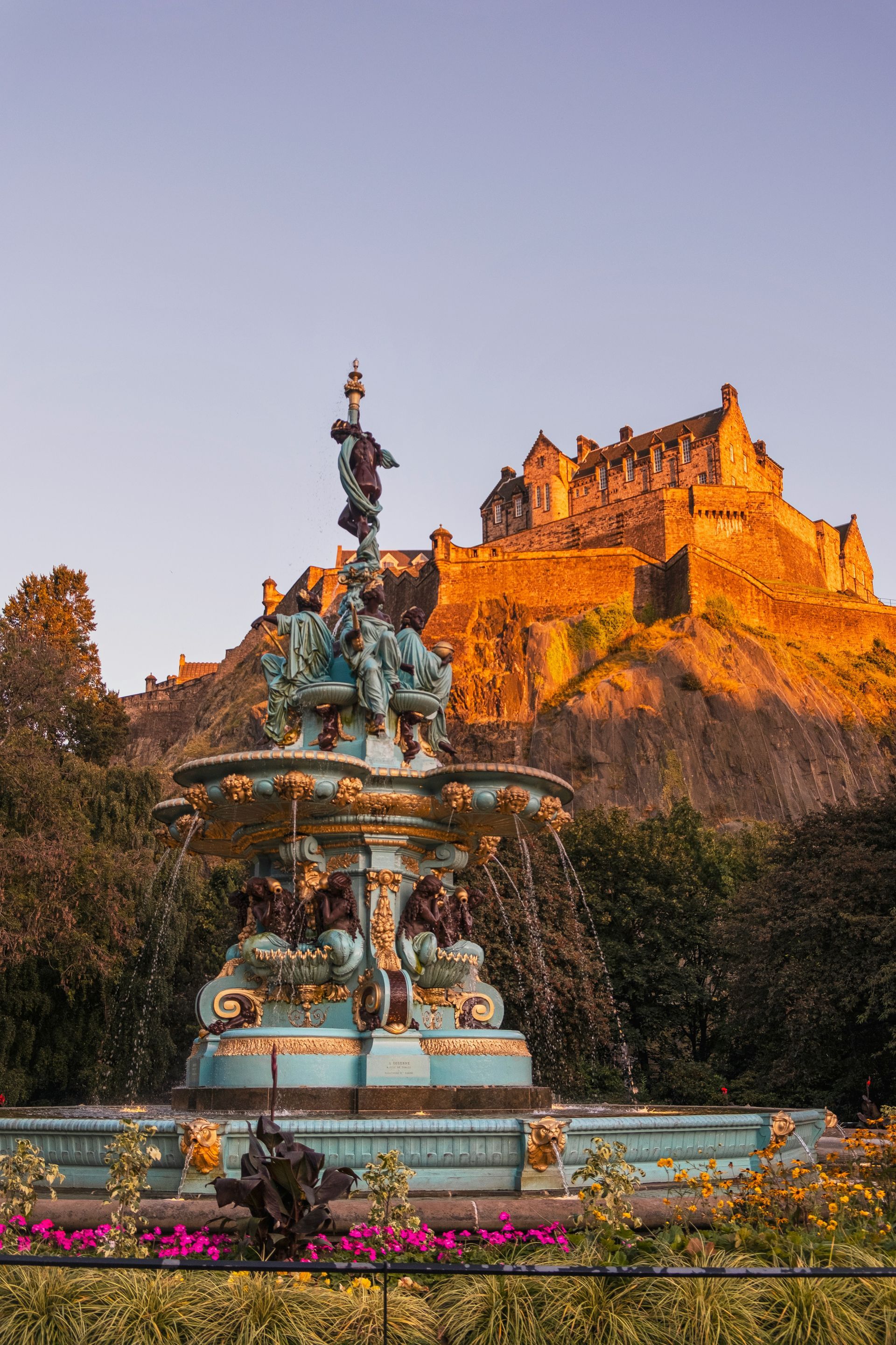 Fountain in foreground, Edinburgh Castle lit by sunset in background. Blue and gold fountain, pink flowers.