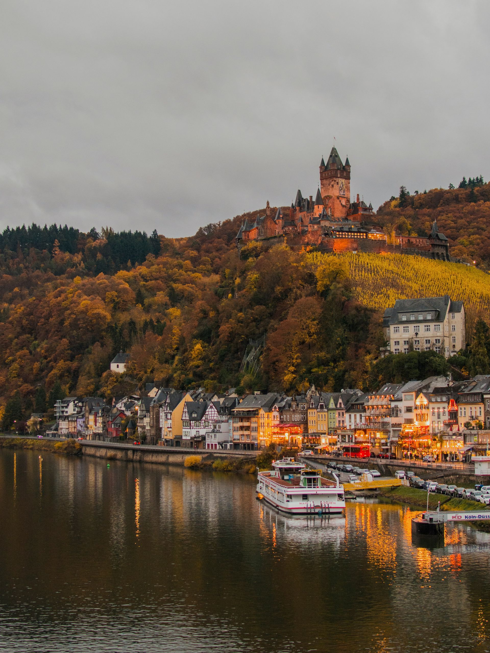 Town of Cochem along a river, with castle on a hill. Colorful buildings, trees in autumn colors, boat on water in Germany.