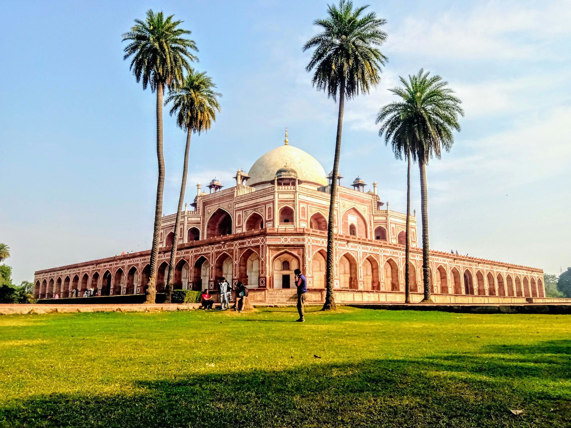 Tomb with large dome, red sandstone facade, surrounded by palm trees and green lawn in New Delhi, India.