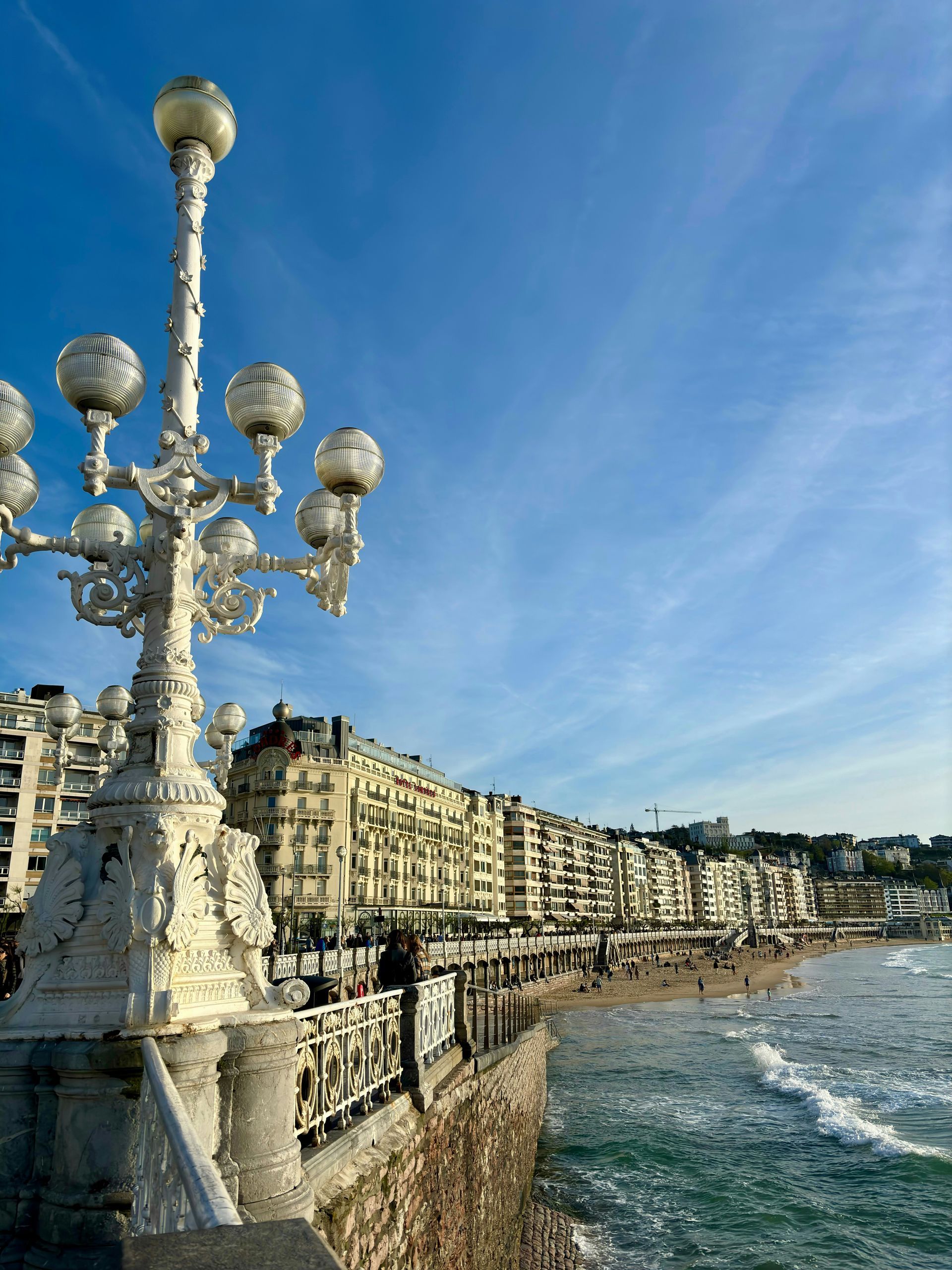 White ornate lamp post on a seaside promenade; beach and buildings in the background in San Sebastián, Spain.