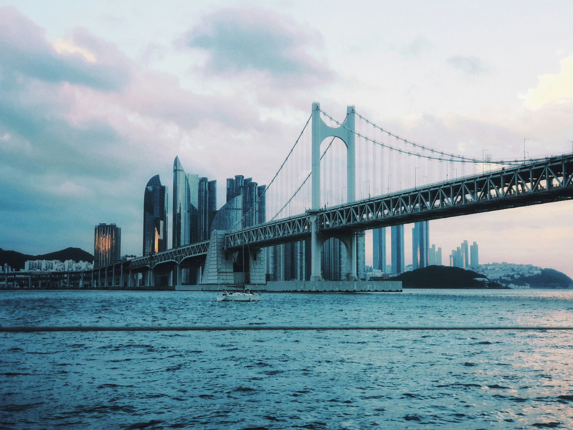 A suspension bridge with skyscrapers in the background at dusk over a body of water in Busan, South Korea.