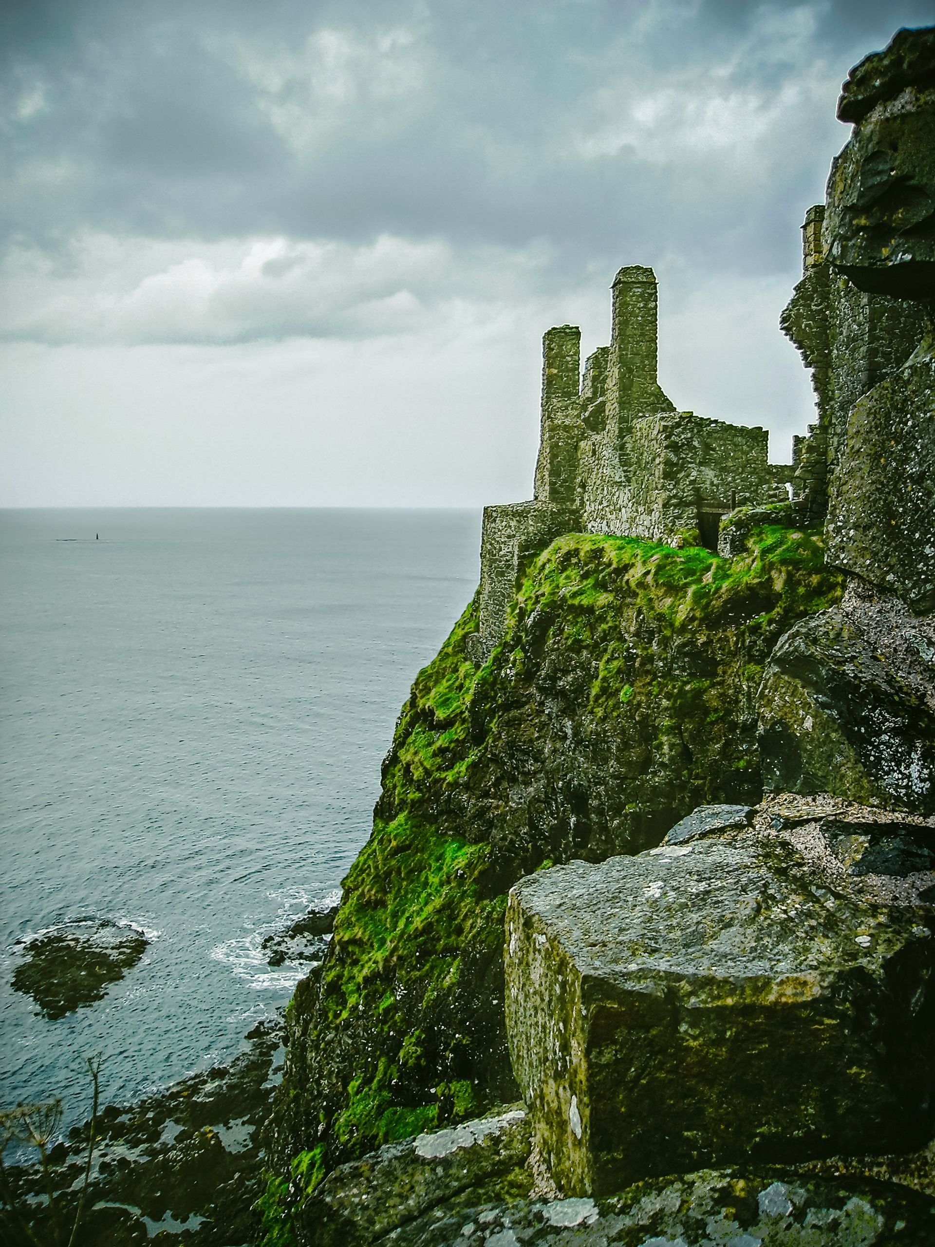 Stone ruins of Dunluce Castle on a green cliff overlooking the ocean under a cloudy sky in Northern Ireland.