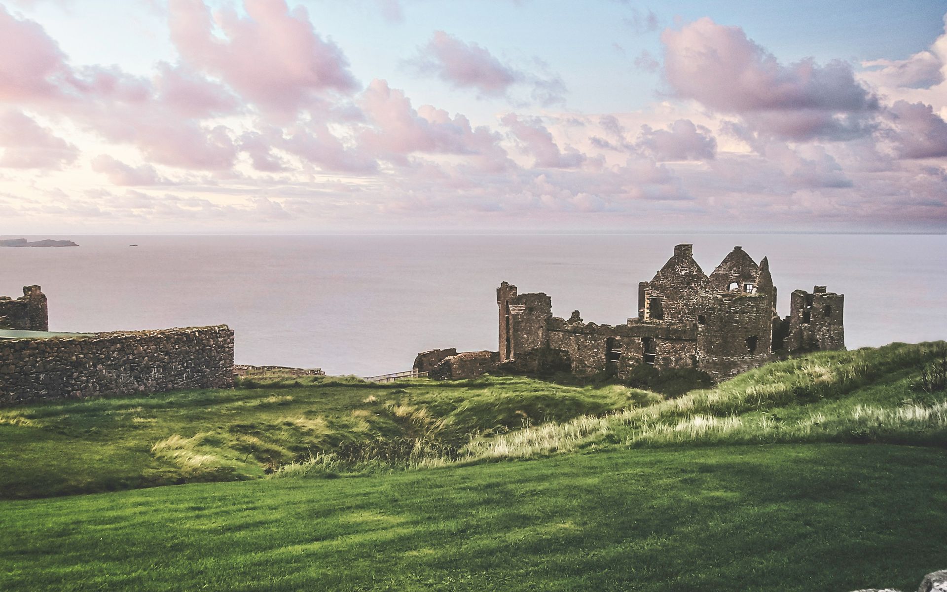 Ruins of Dunluce Castle on a grassy cliff, overlooking a calm sea under a pastel sky in Northern Ireland.