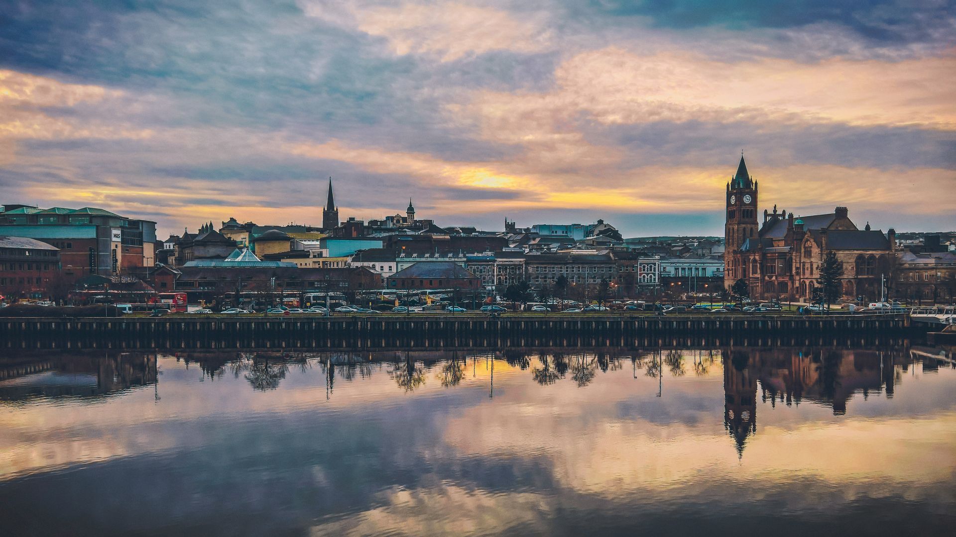 City skyline at dusk reflected in still water, with orange and blue sky in Londonderry, Northern Ireland.
