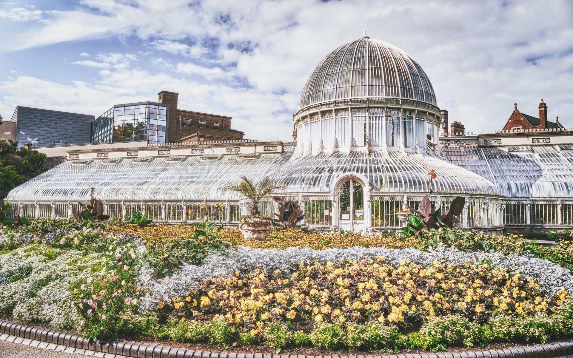 Botanical garden with glass greenhouse and colorful flowers under a cloudy sky in Belfast, Northern Ireland.