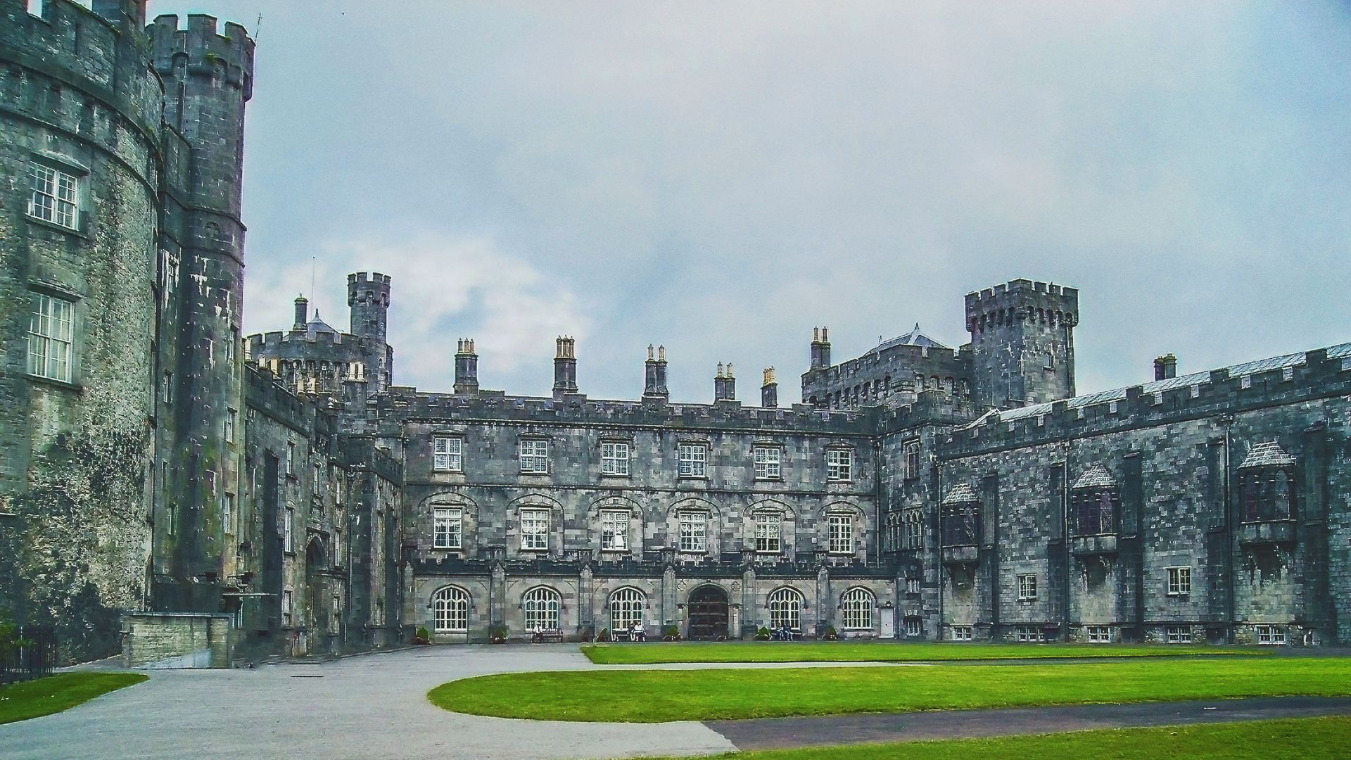 Exterior view of Kilkenny Castle in Ireland, gray stone, overcast sky, green lawn.