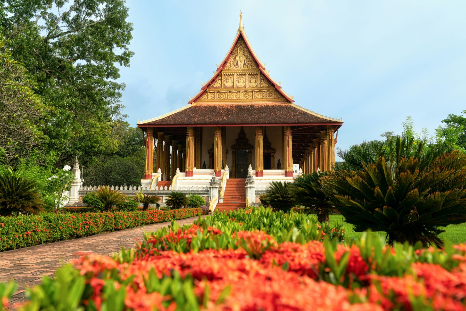 Temple in Vientiane, Laos. Red flowers and a path lead to the beige and gold structure with a tall roof under a blue sky.