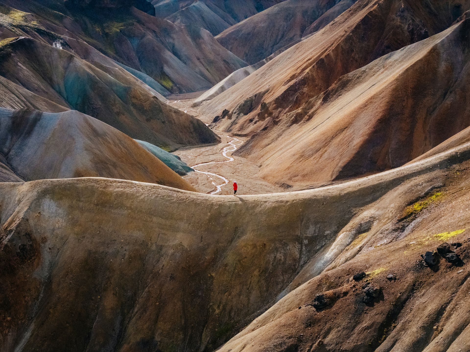 Hiker in red jacket stands on a ridge, overlooking colorful mountains and valleys in Landmannalaugar, Iceland.