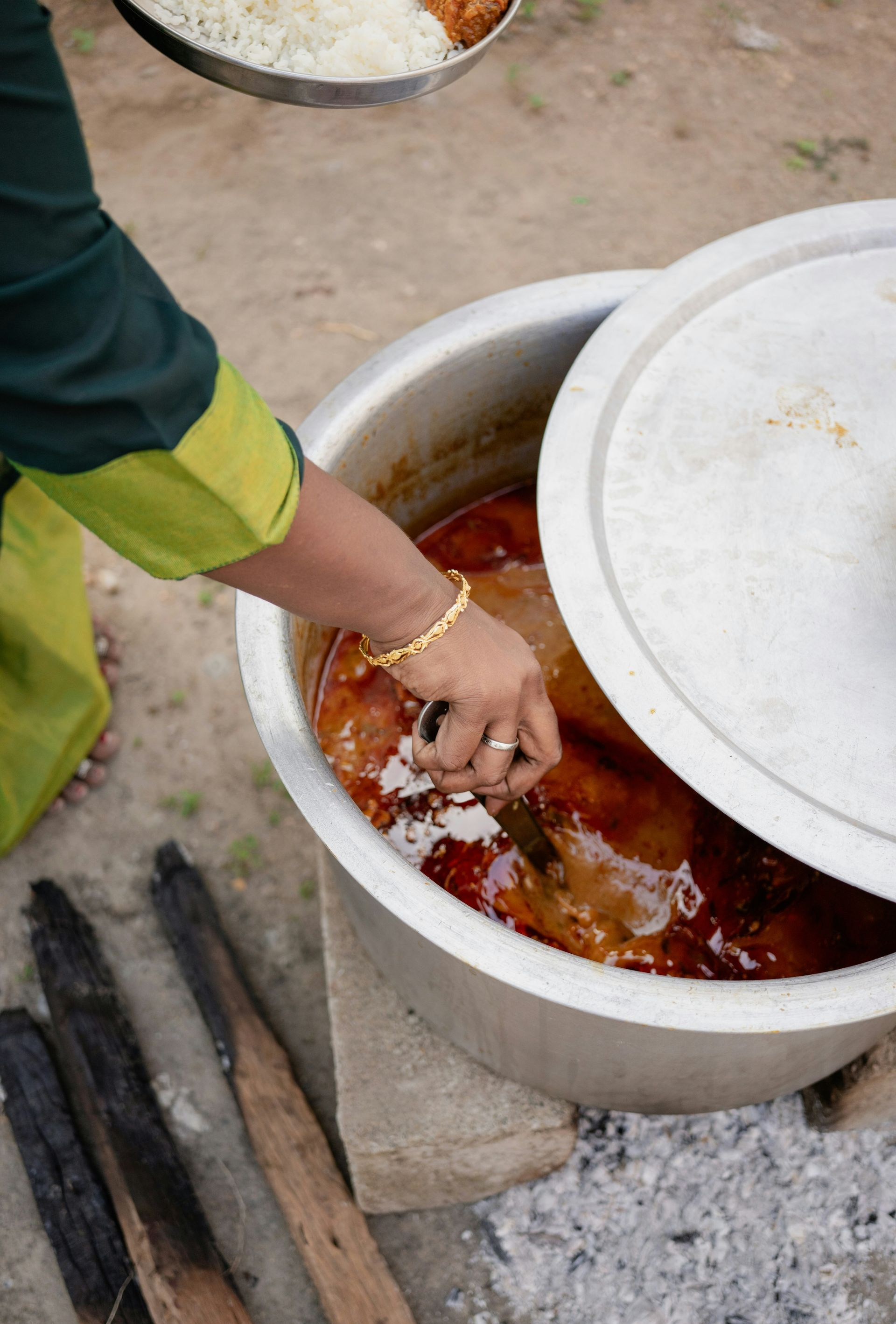 Woman reaching into a large pot of food outdoors, with a plate of food in the background in India.