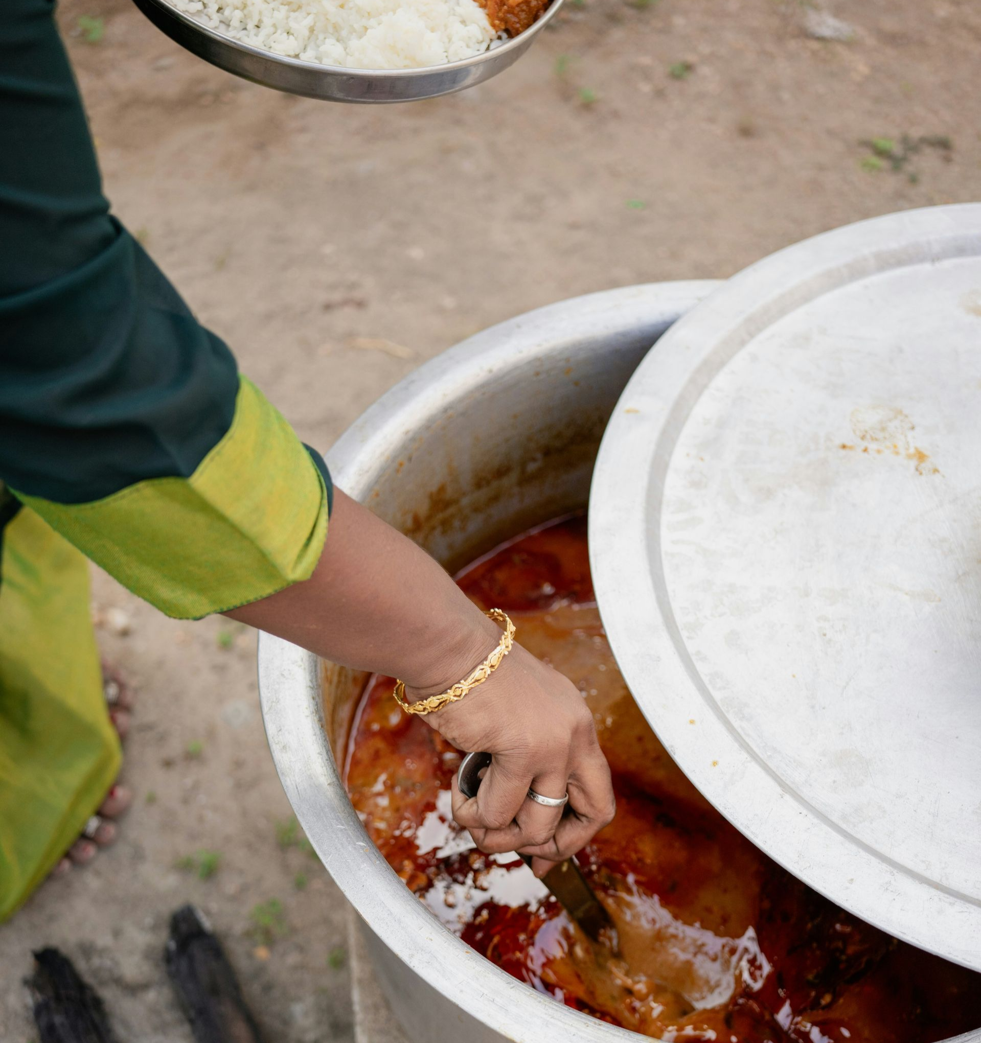 Woman reaching into a large pot of food outdoors, with a plate of food in the background in India.