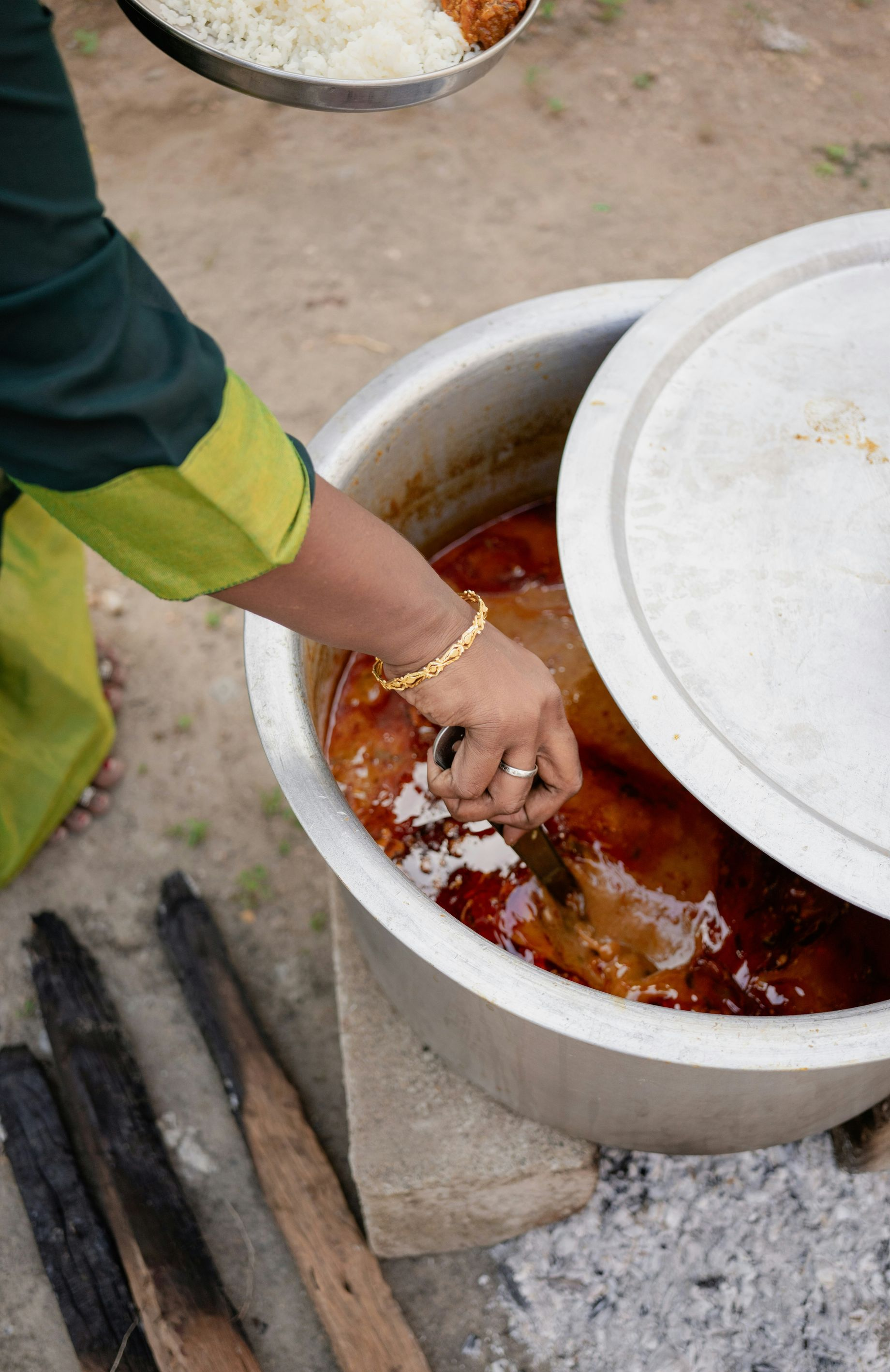 Woman reaching into a large pot of food outdoors, with a plate of food in the background in India.
