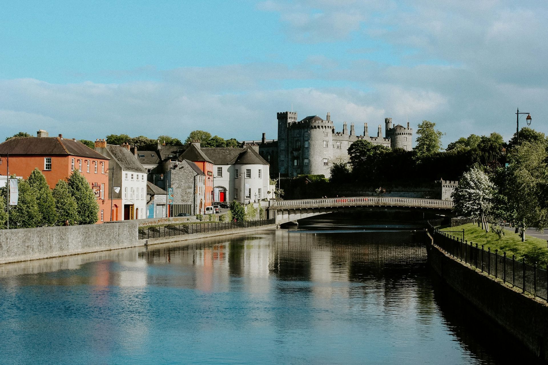 River view of Kilkenny Castle in Ireland with buildings along the water and a bridge in Ireland.