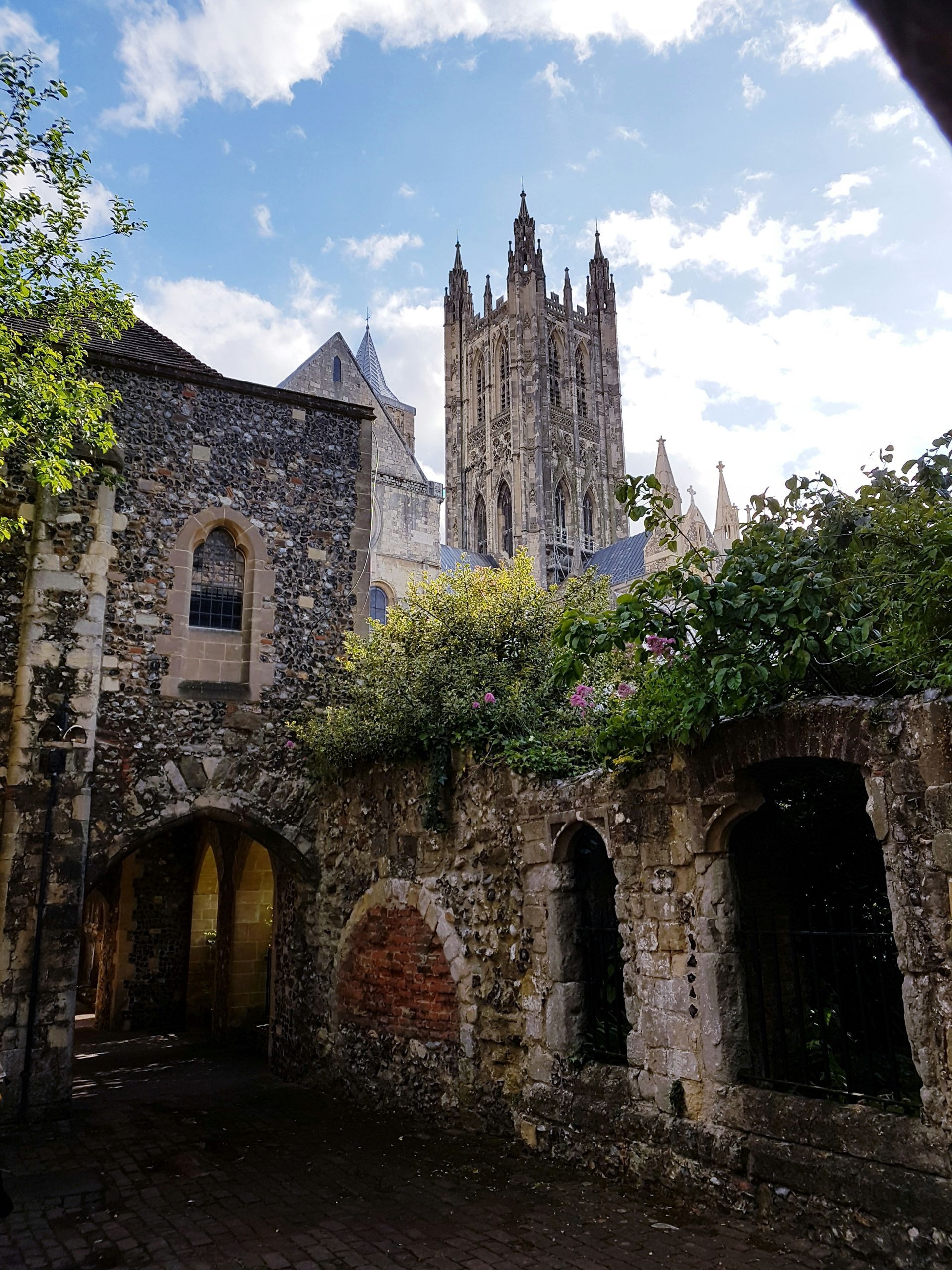 Old stone buildings and arched openings with Canterbury Cathedral in the background, cloudy sky in England.