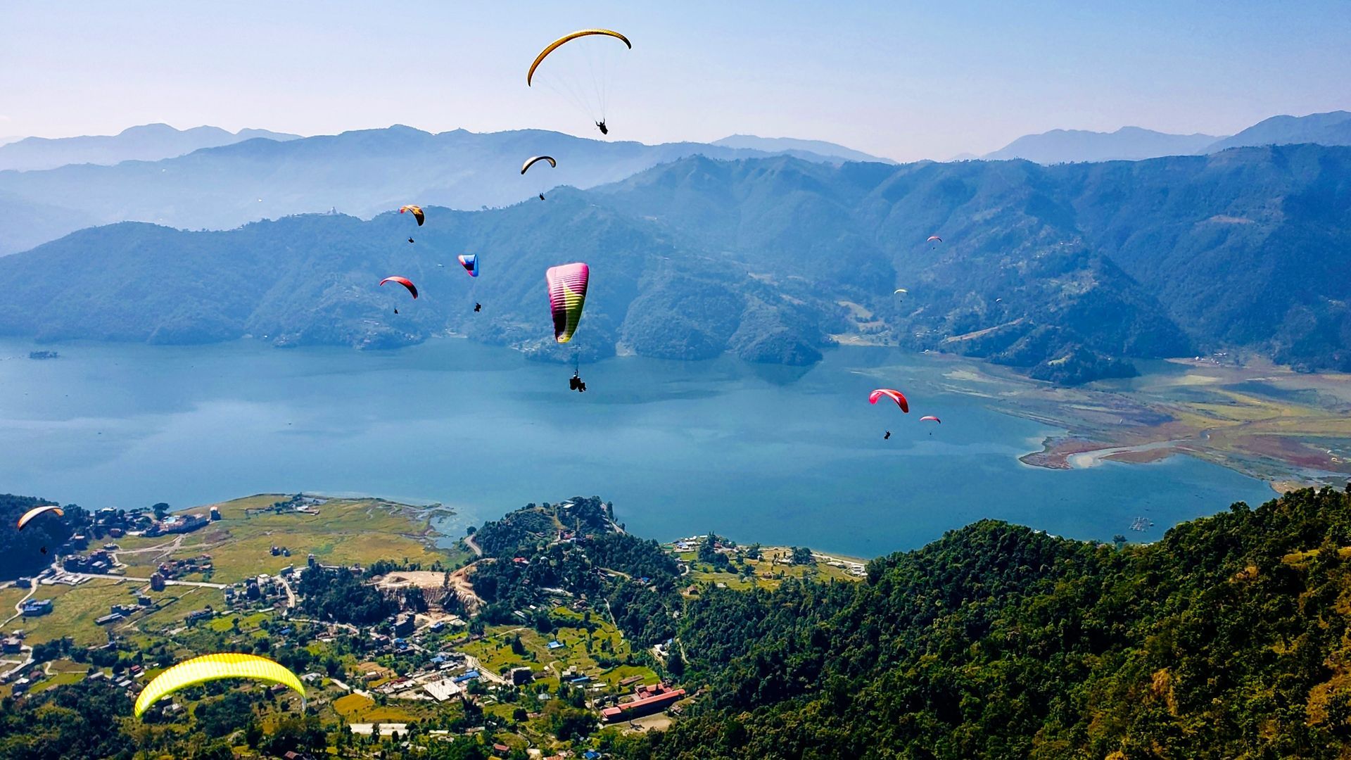 Paragliders soar over Phewa Lake in Nepal, with mountains and forests in the background.