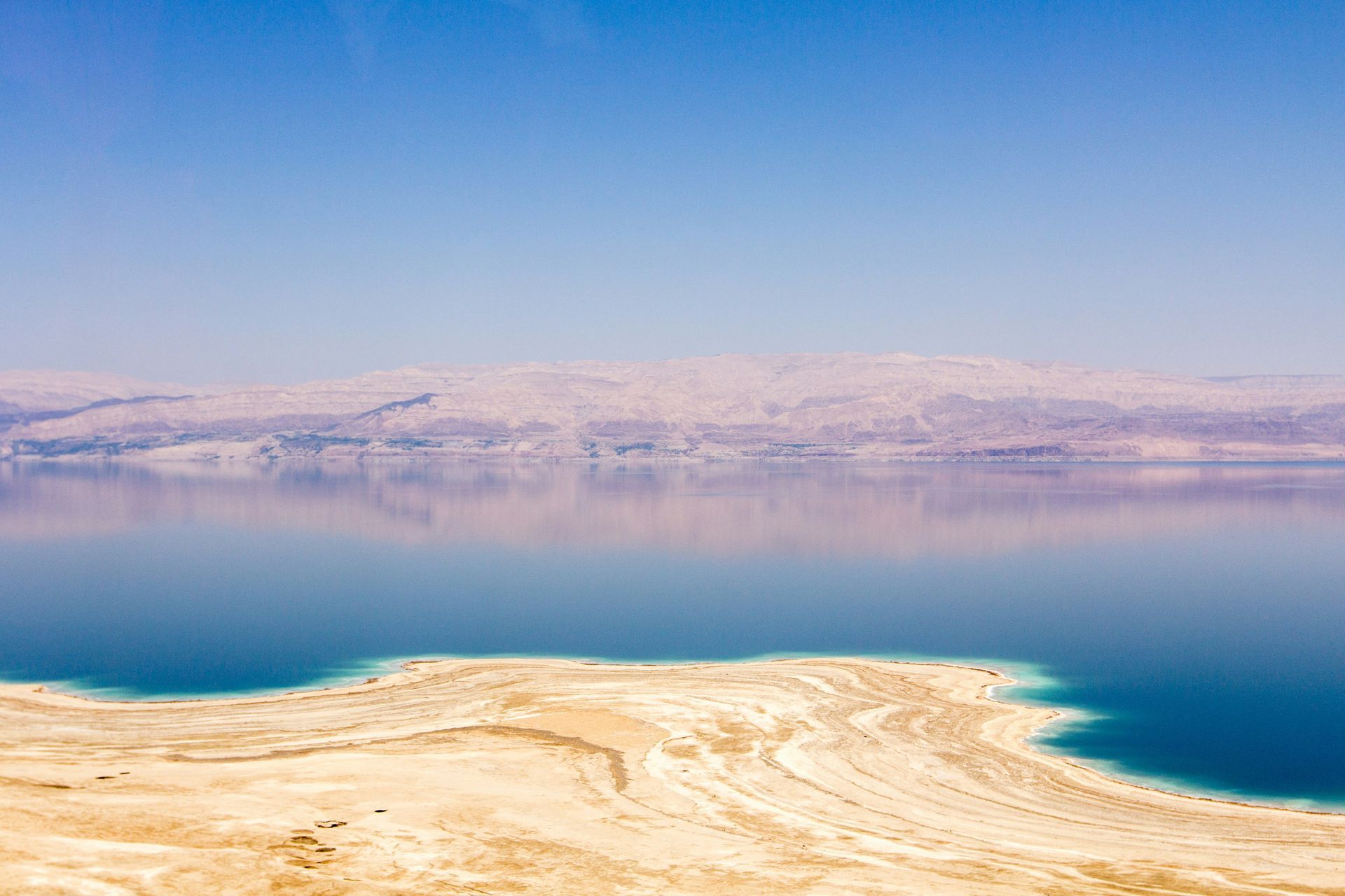Dead Sea with a tan beach, reflective water, and hazy mountains under a blue sky in Jordan.