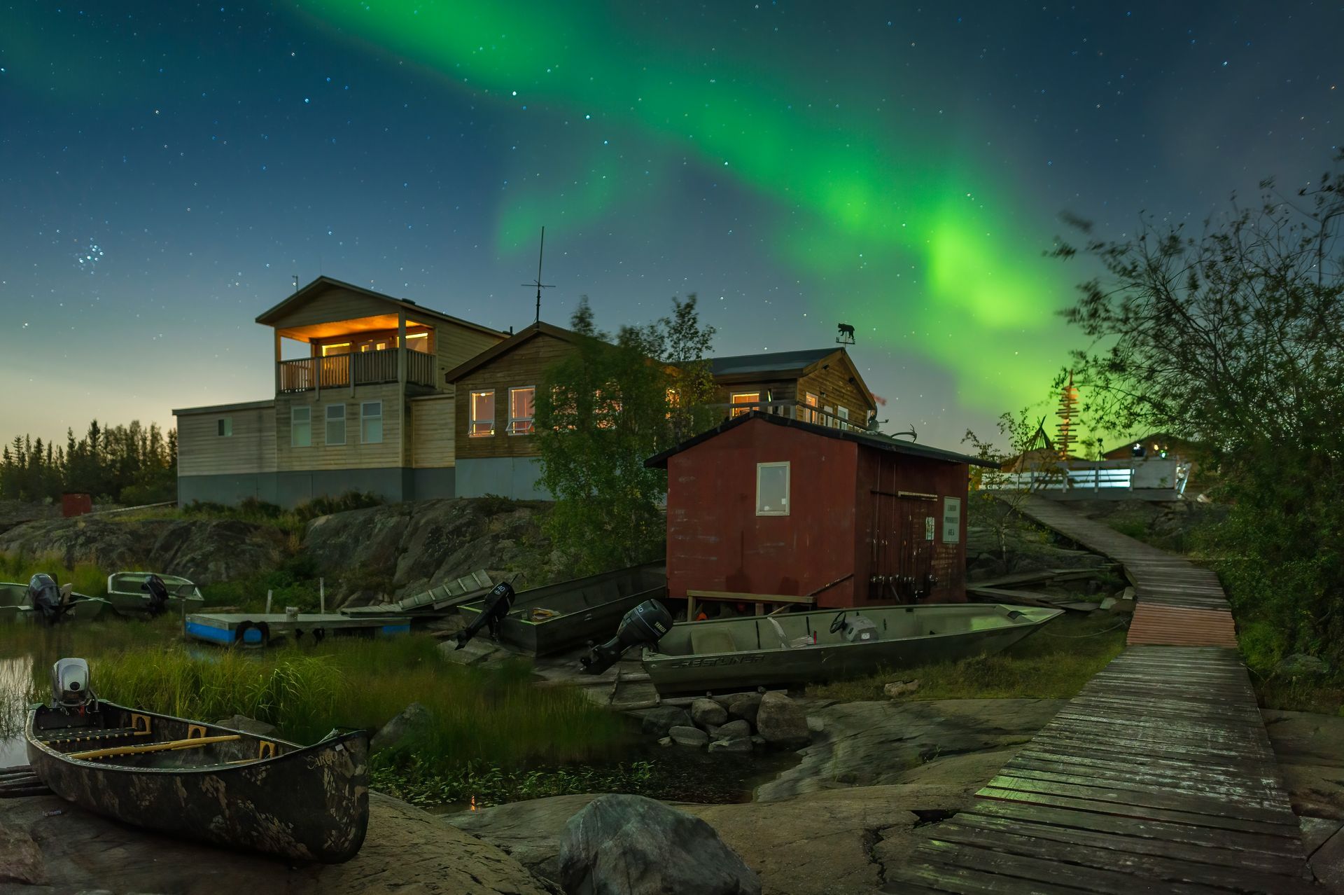 Northern Lights illuminate cabins on a lake shore at night. A small boat rests in the foreground in Yellowknife, Canada.