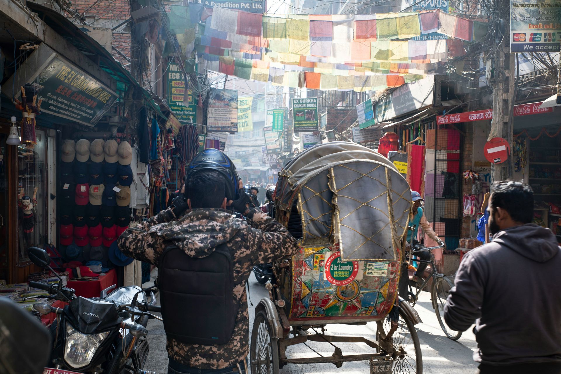 Busy street scene in Kathmandu, Nepal, with a rickshaw, people, shops, and colorful prayer flags overhead.